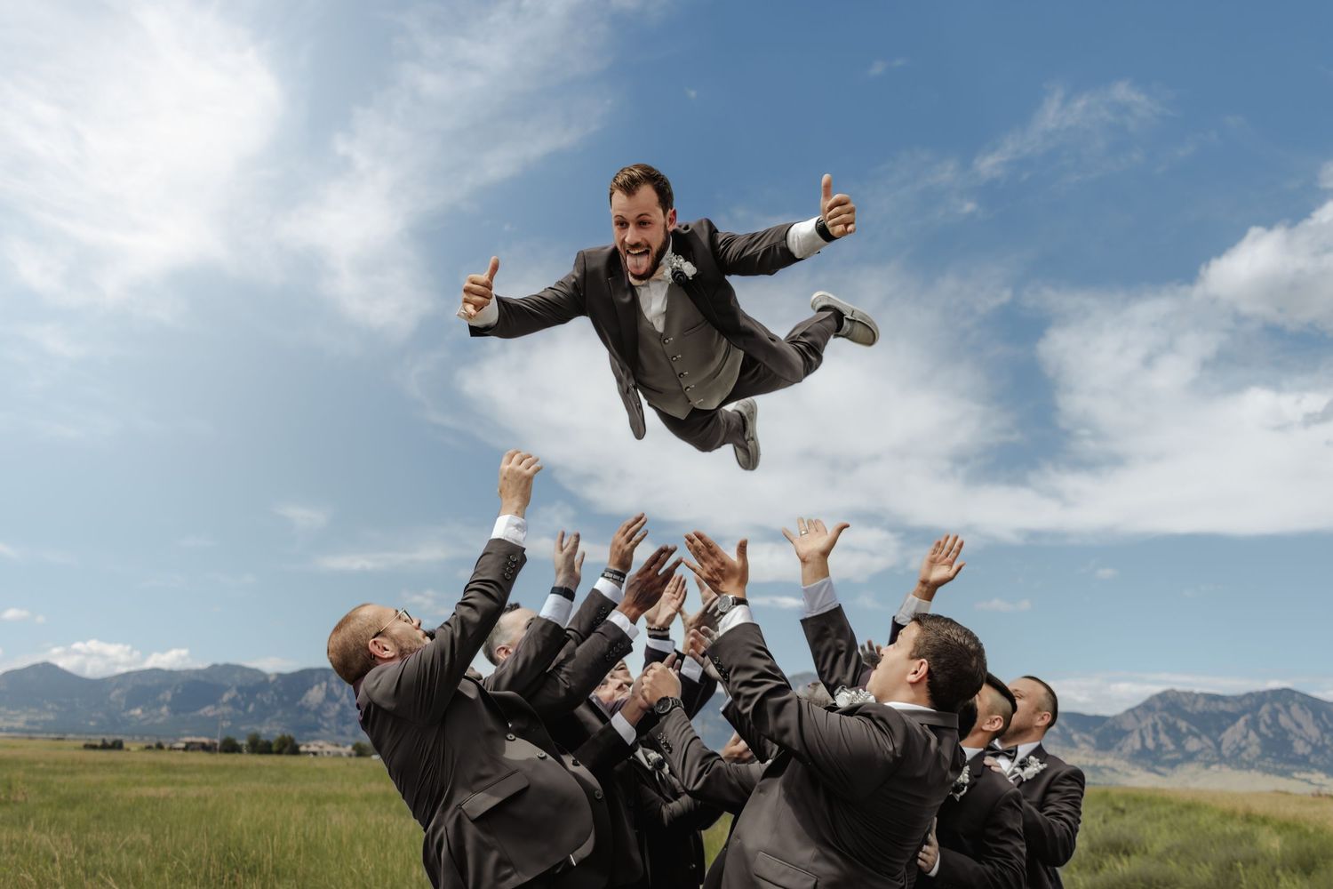 Groomsmen tossing a celebrating groom into the air in an open field under a blue sky.