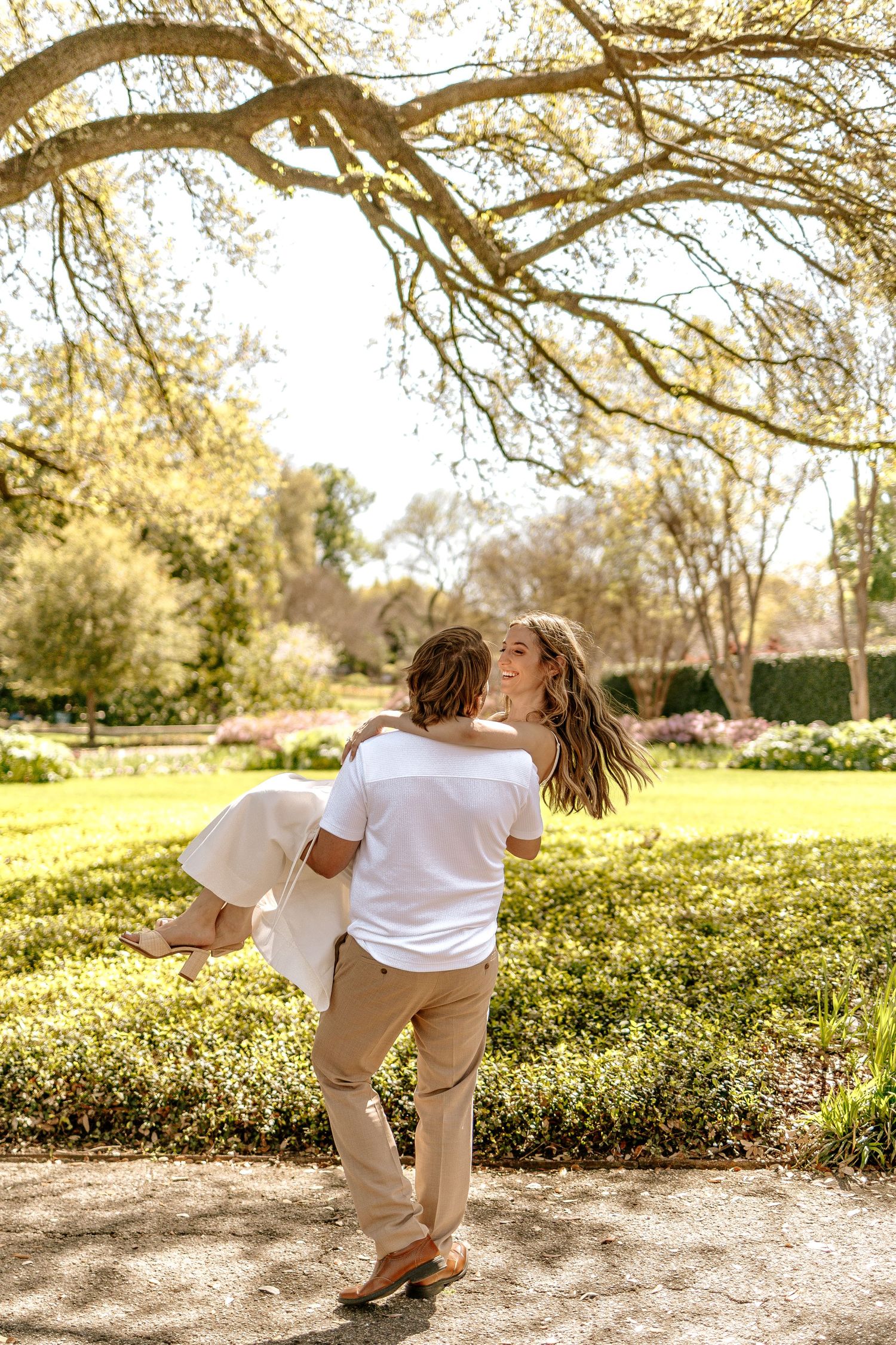 Man lifting and spinning woman in a lush green park under a large tree during engagement photos.
