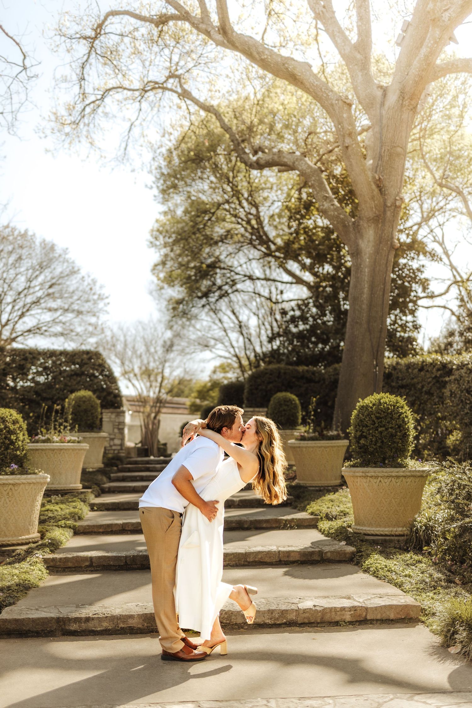 Couple sharing a romantic kiss on sunlit garden pathway lined with topiaries and large trees.