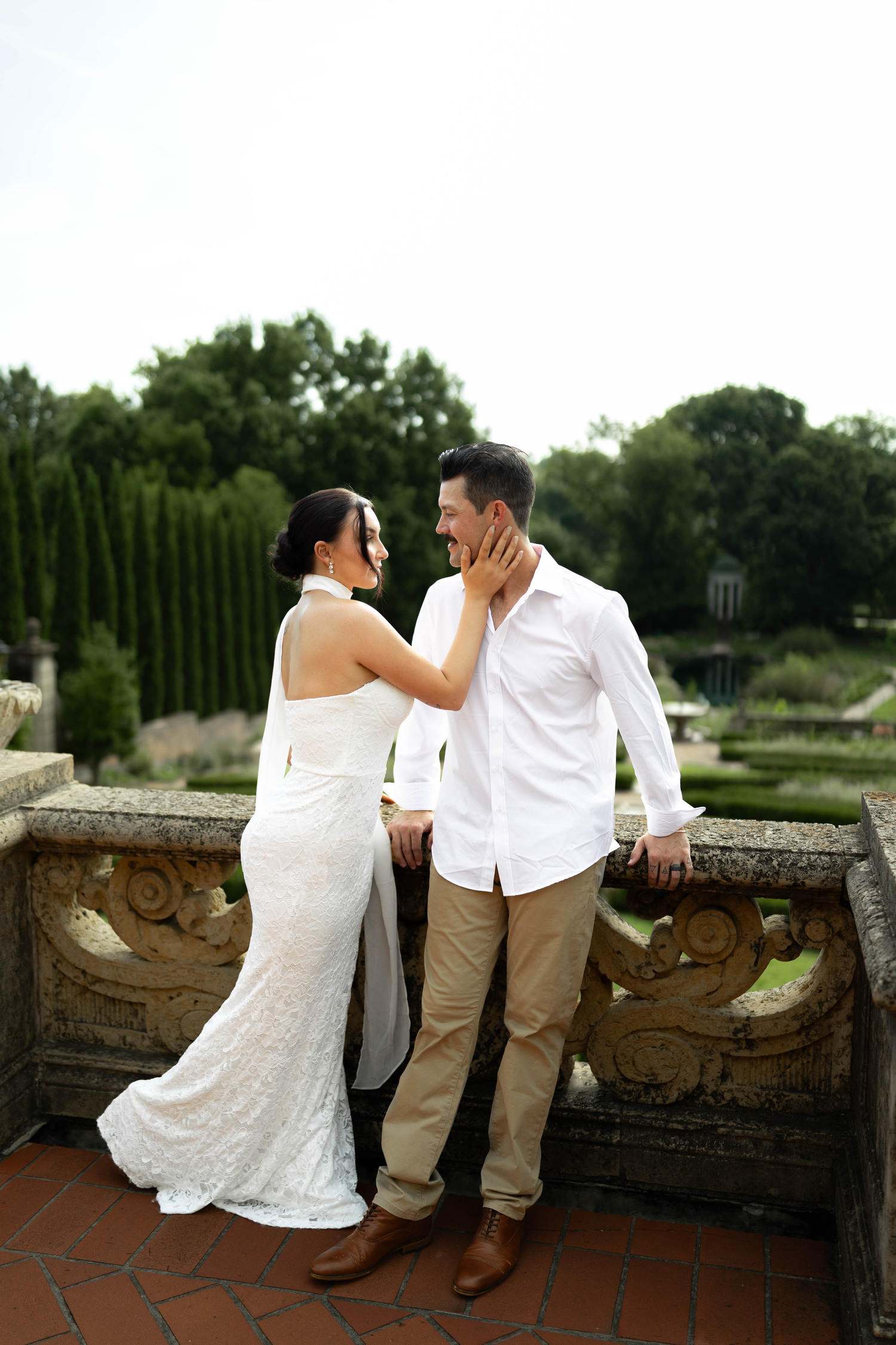 Elegant couple in wedding attire sharing a romantic moment on a stone balcony surrounded by lush gardens.