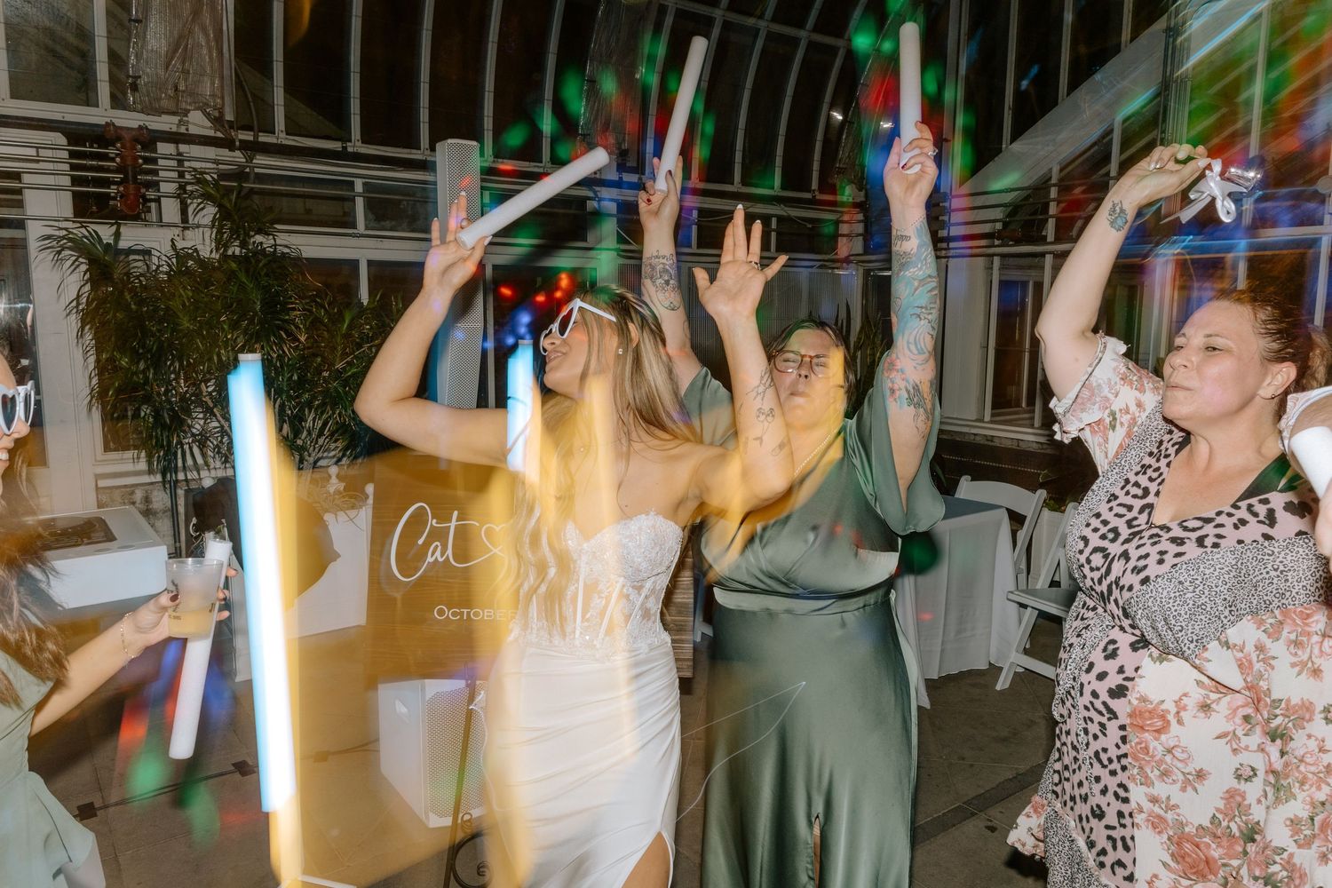 Bride and guests dancing with hands raised under colorful lights at a wedding reception.