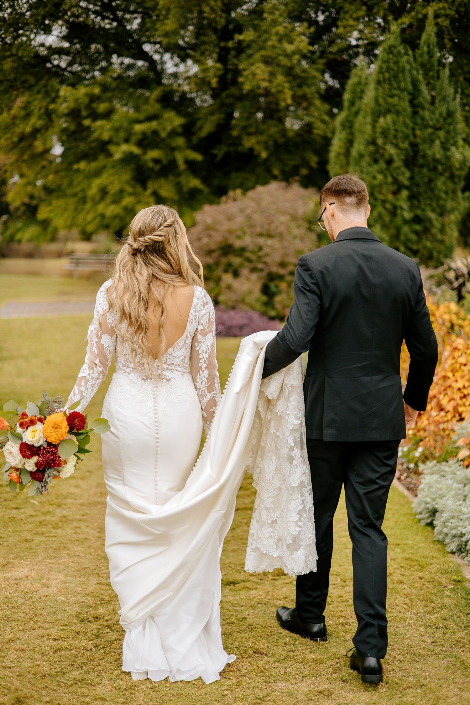 Groom in black suit carries bride's lace train as couple walks through a vibrant autumn garden holding a floral bouquet.