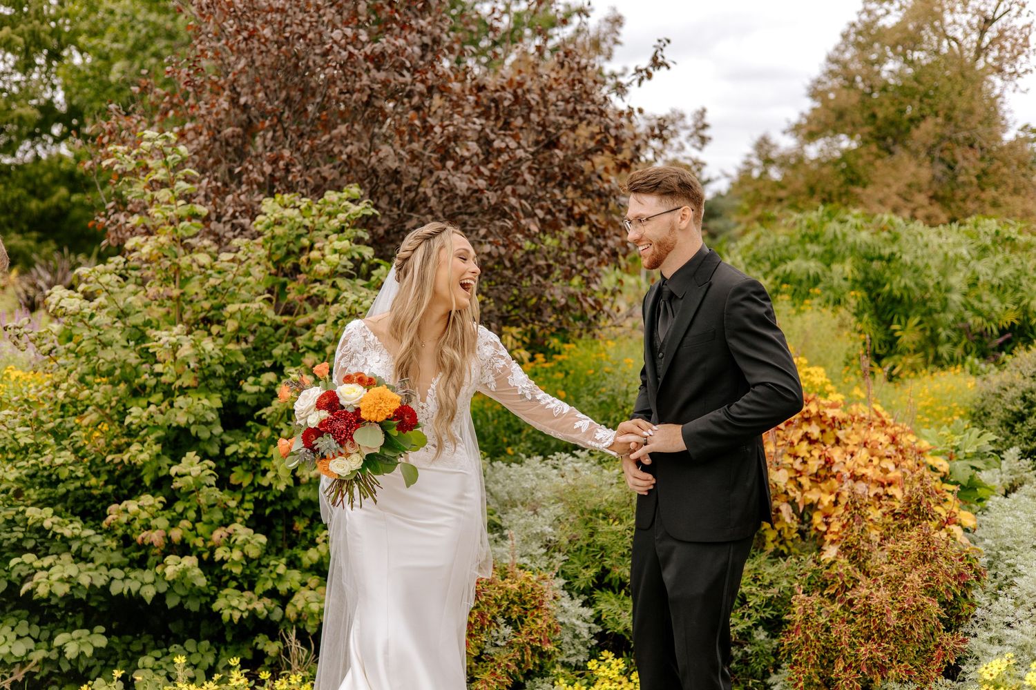 Bride in white gown holding colorful bouquet takes grooms hand in autumn garden wedding photo.