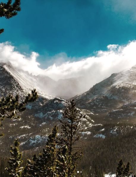 Snow-dusted Rocky Mountain peaks with evergreen trees and dramatic clouds against a vivid blue sky.
