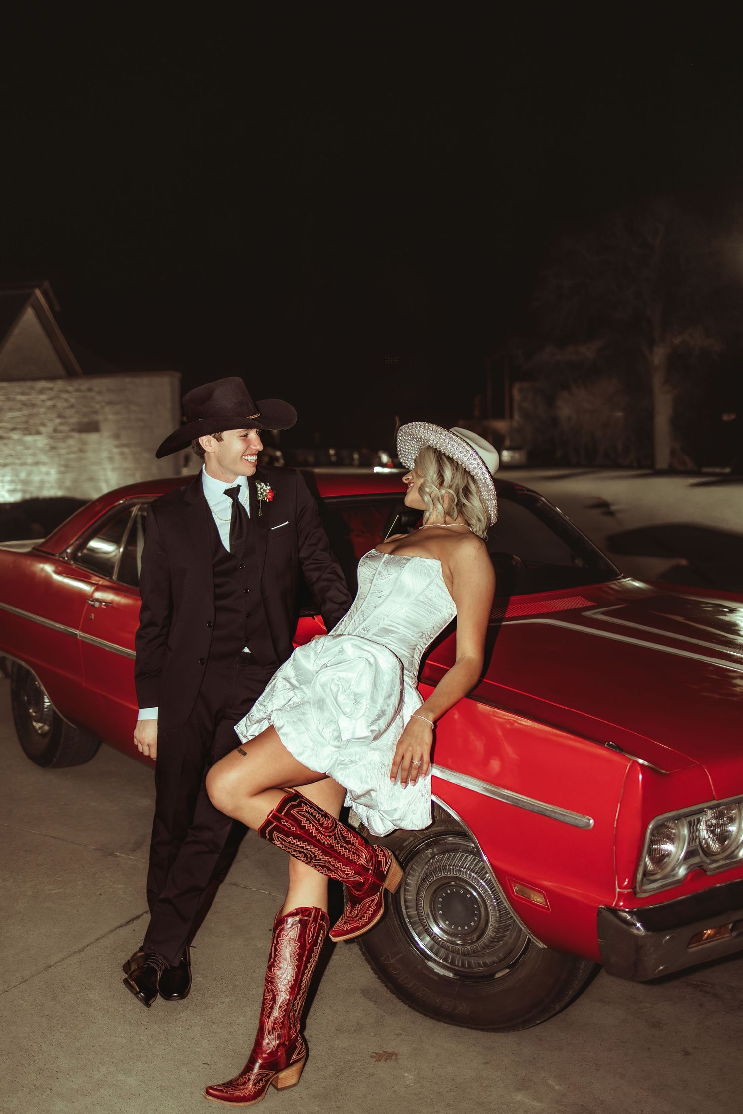 Couple in western attire pose by red classic car at night, bride in white dress and red cowboy boots.
