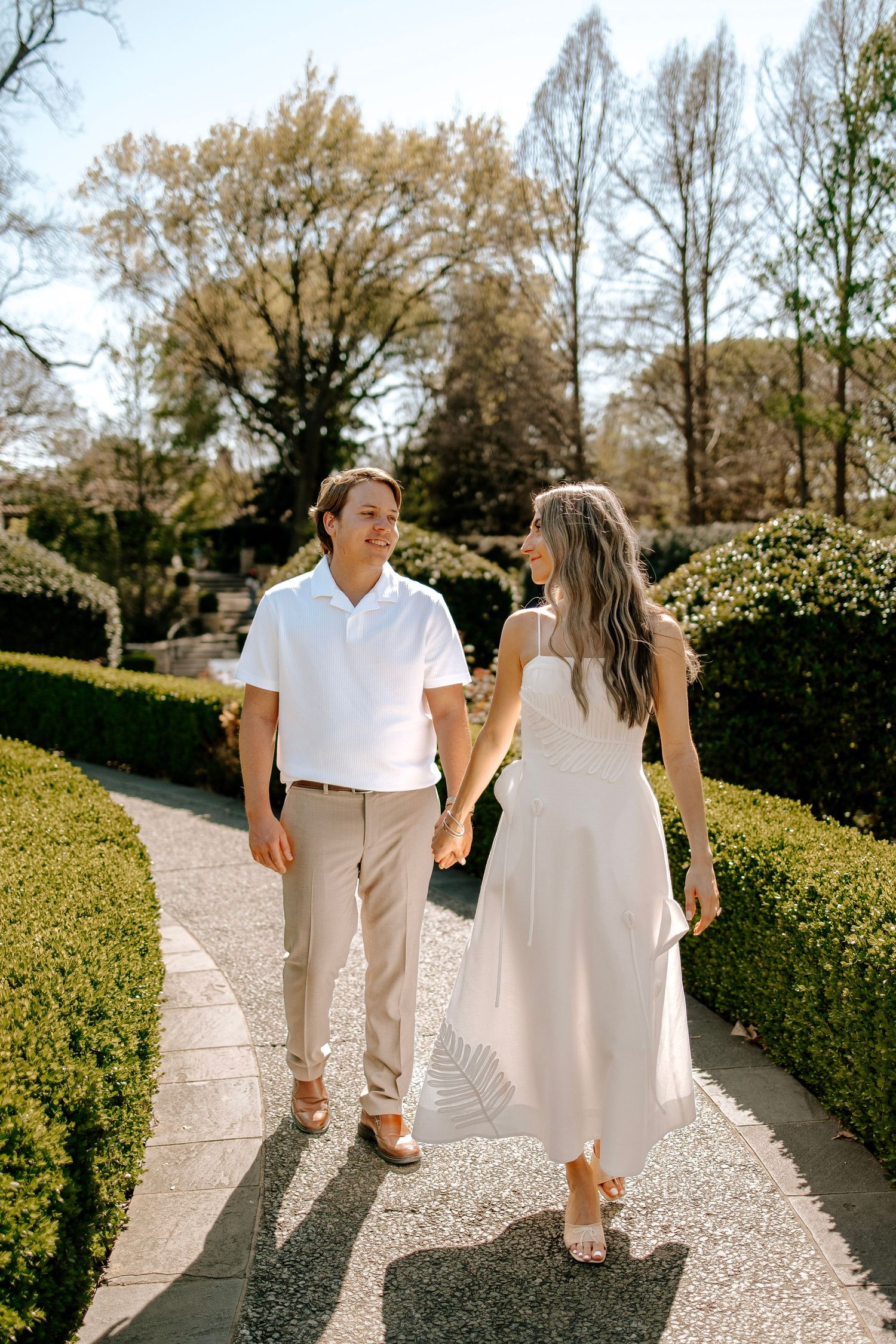 Couple walking hand in hand along a manicured garden path, smiling at each other on a sunny day.