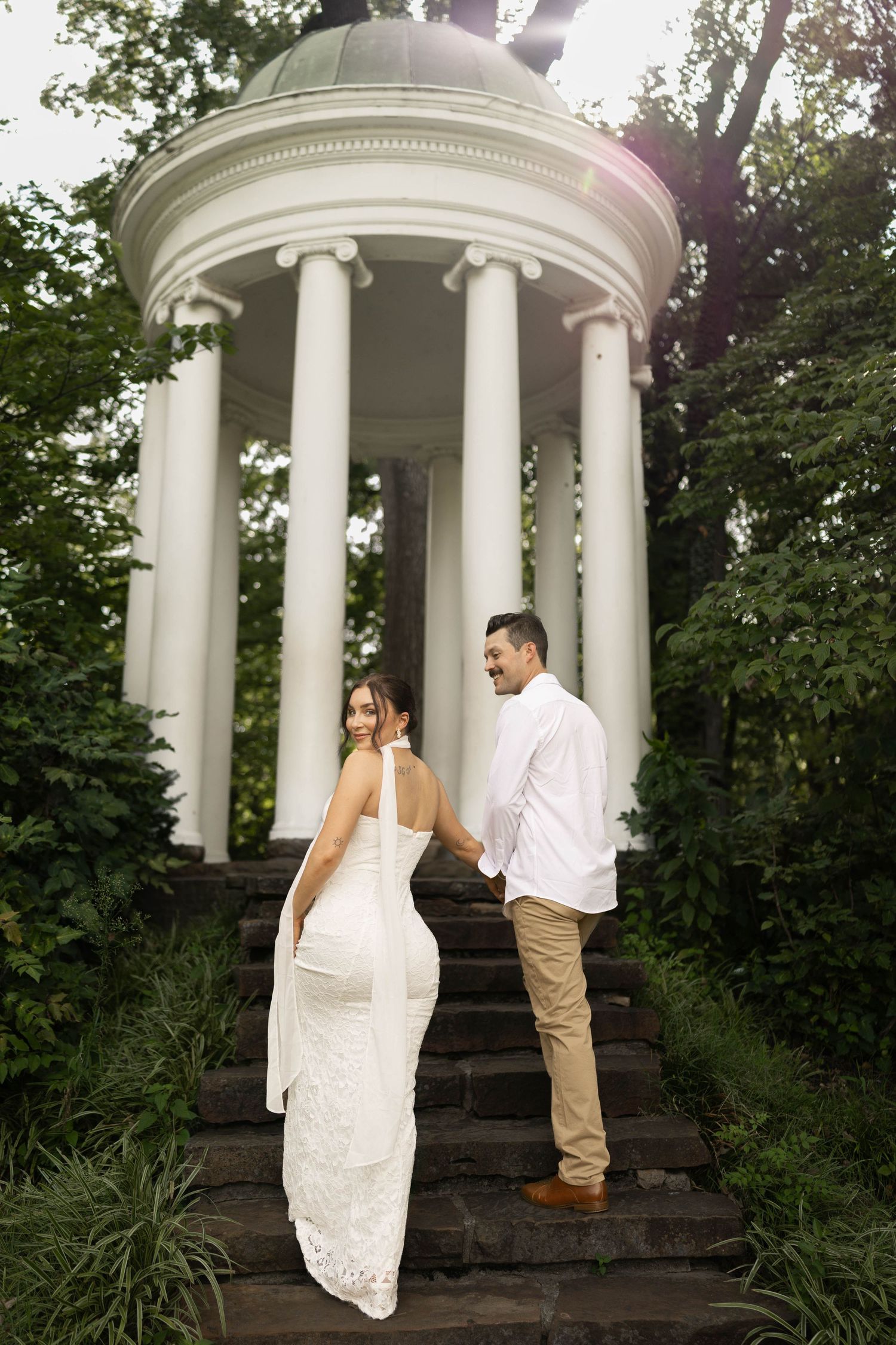 Bride and groom hold hands ascending stone steps toward a white classical gazebo surrounded by trees.