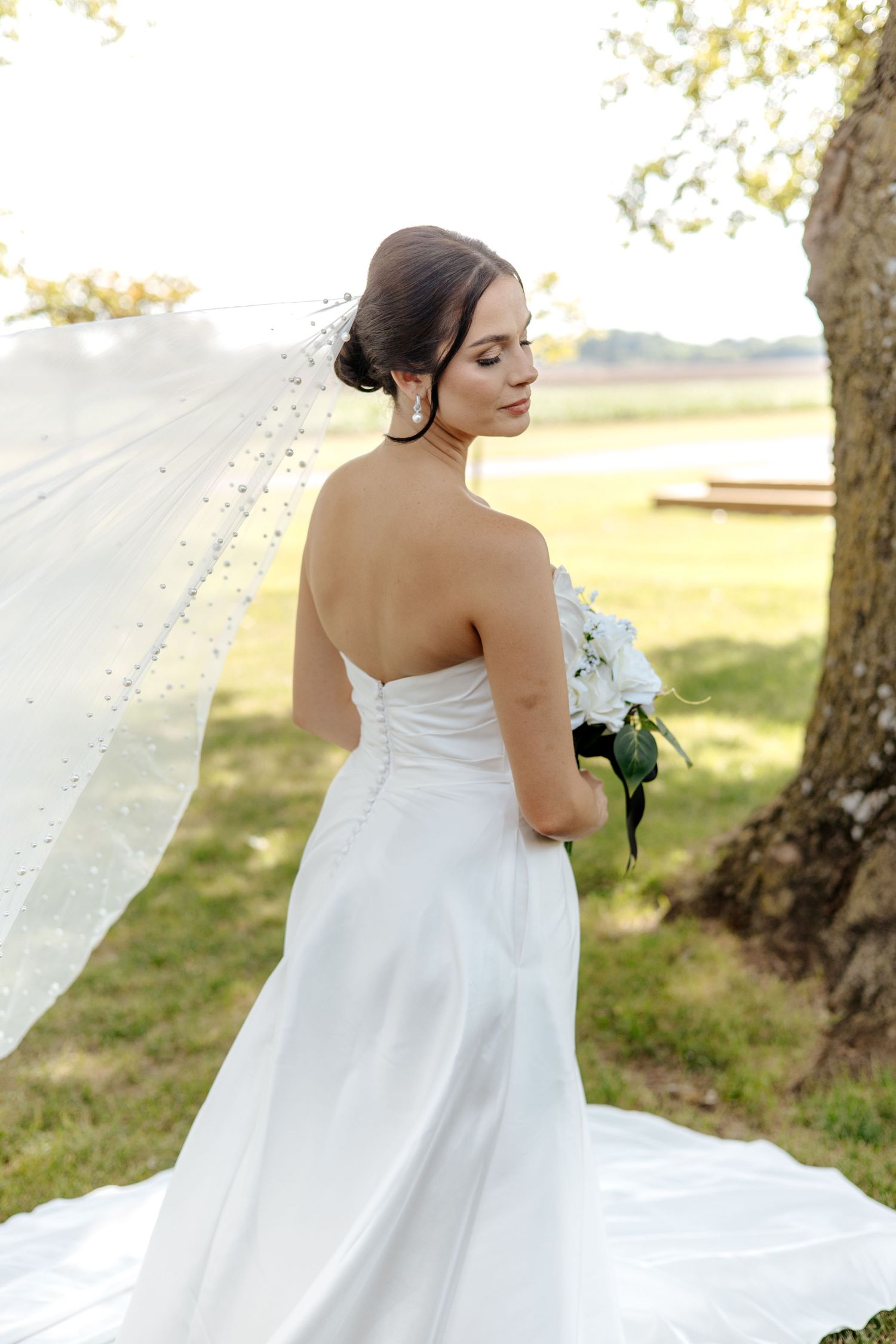 Bride in strapless white gown with flowing veil holds bouquet while standing near tree outdoors.