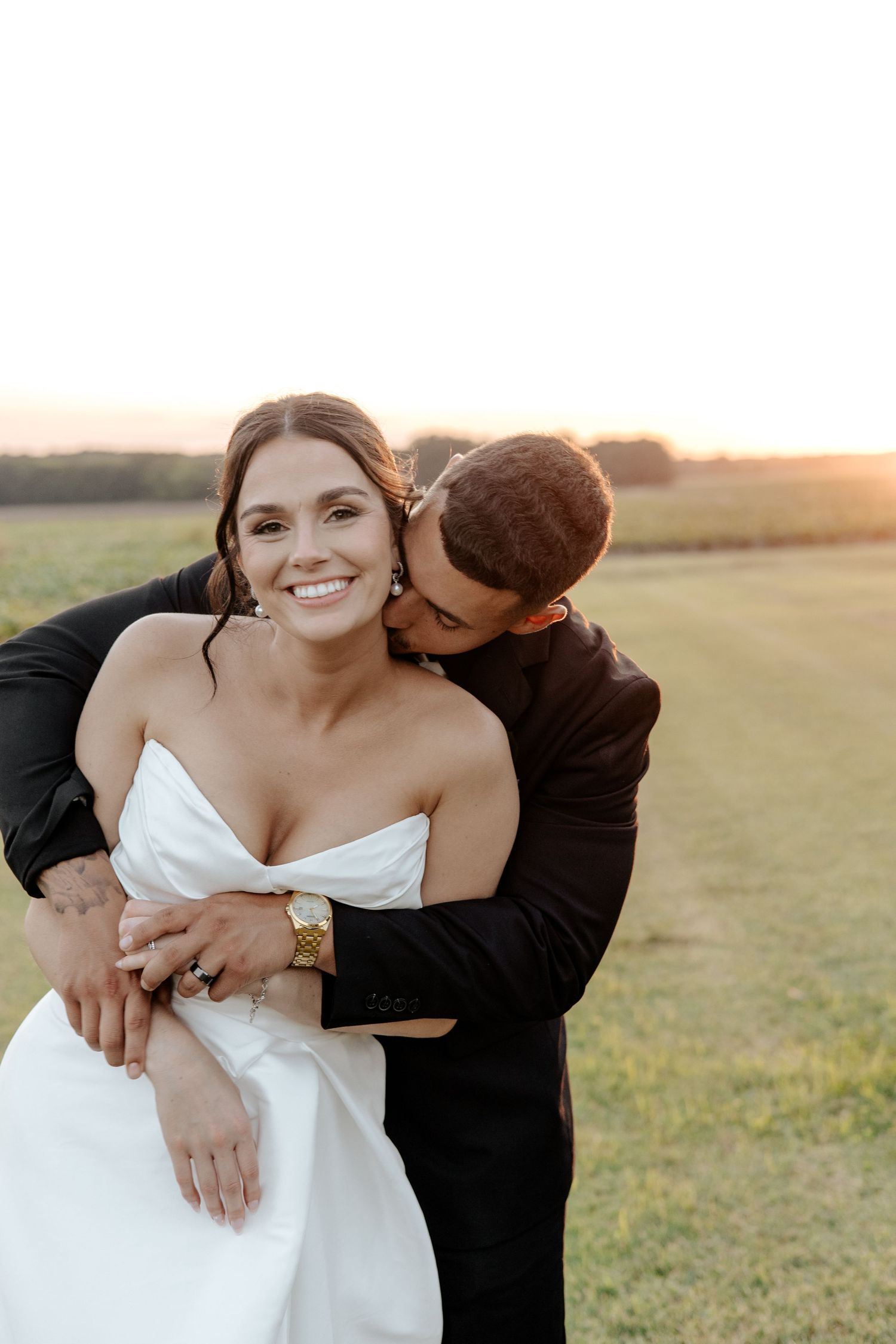 Smiling bride in strapless white gown embraced by groom at sunset in an open field.