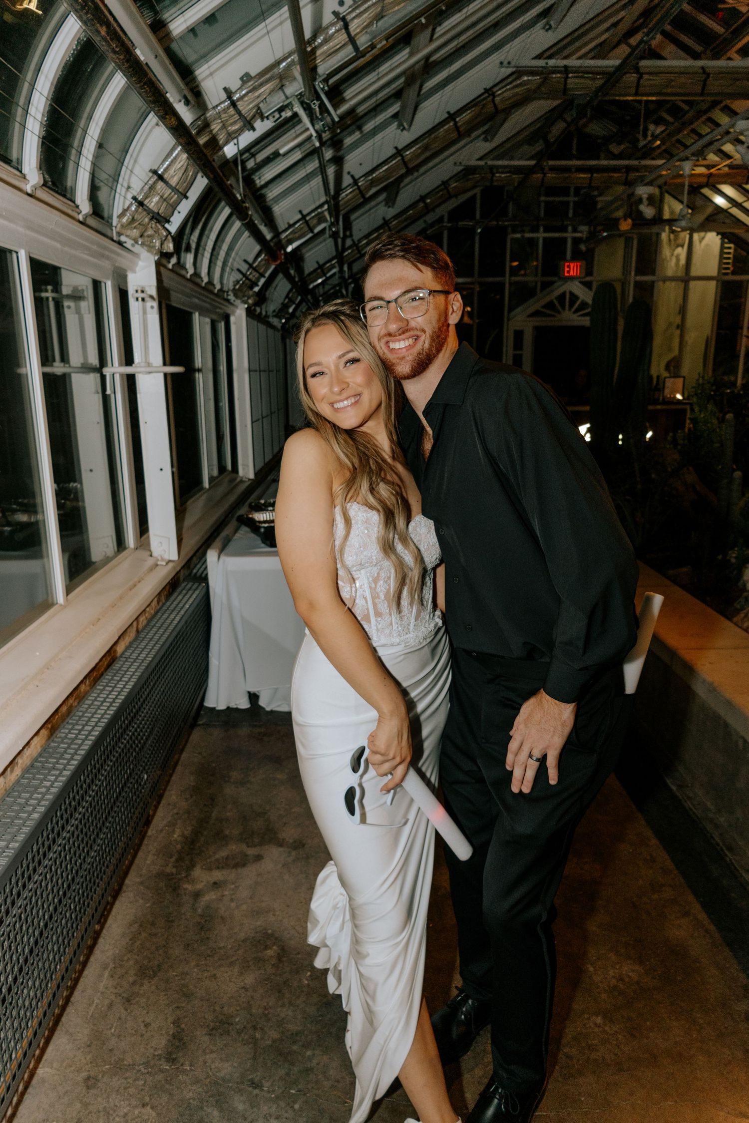 Bride in white gown and groom in black suit posing together at a greenhouse wedding venue.