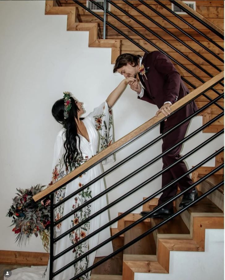 Bride and groom share a romantic moment on modern staircase, bride holding bouquet wearing floral crown, groom in burgundy suit.