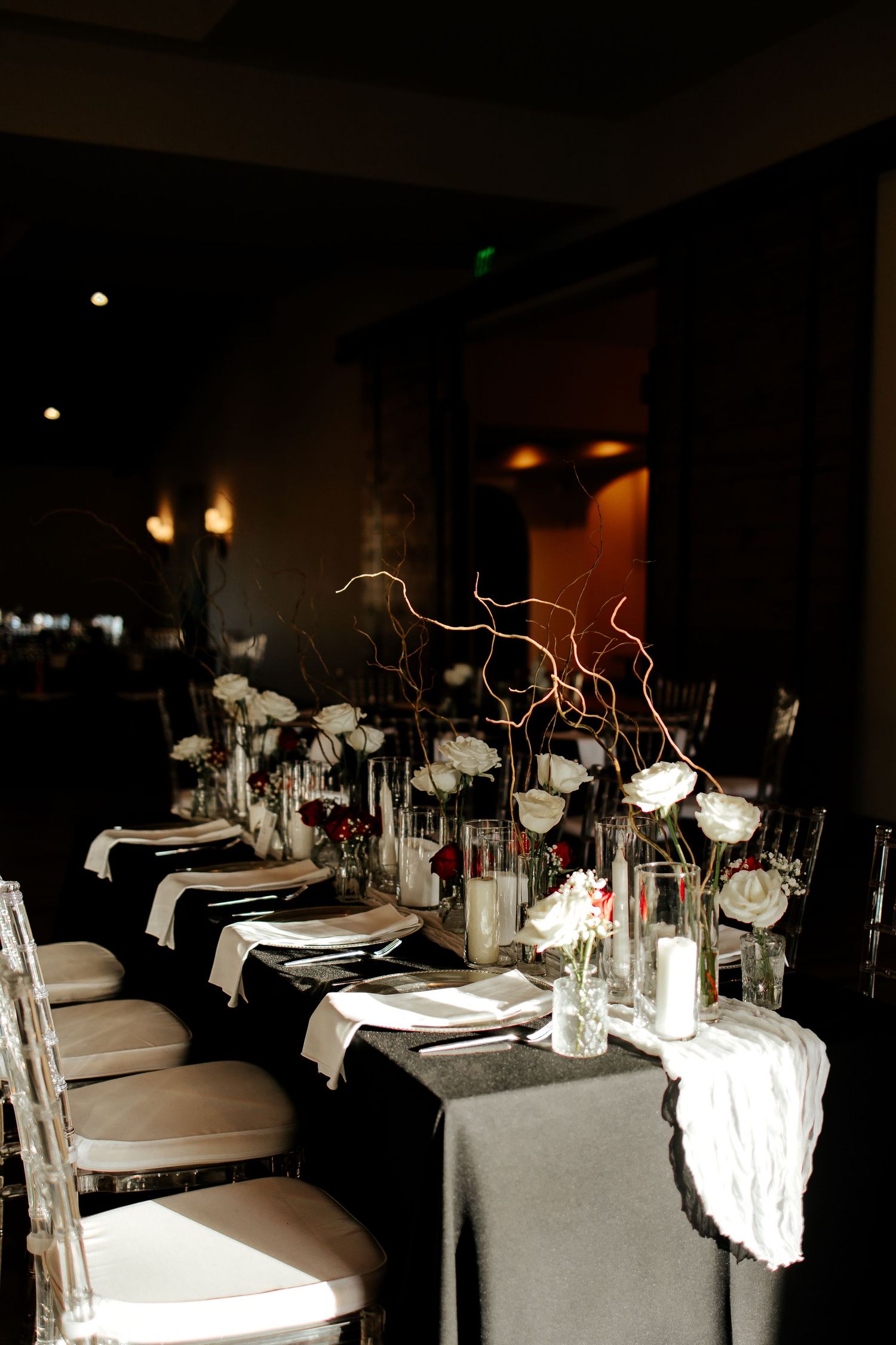 Elegant dark wedding reception table with white roses, candles, and twisted branch centerpieces.