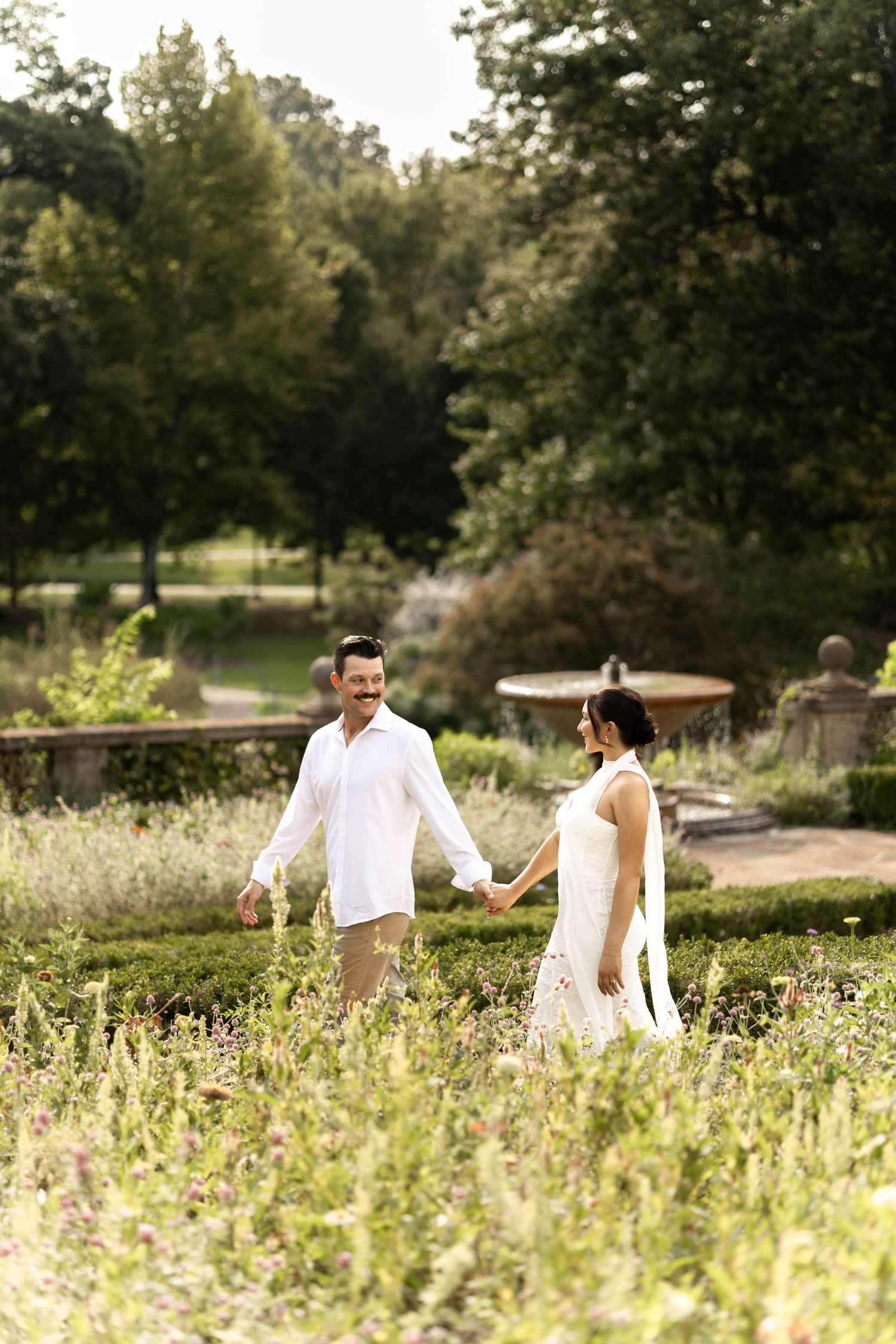Couple in white outfits holding hands while walking through a lush garden park on a sunny day.