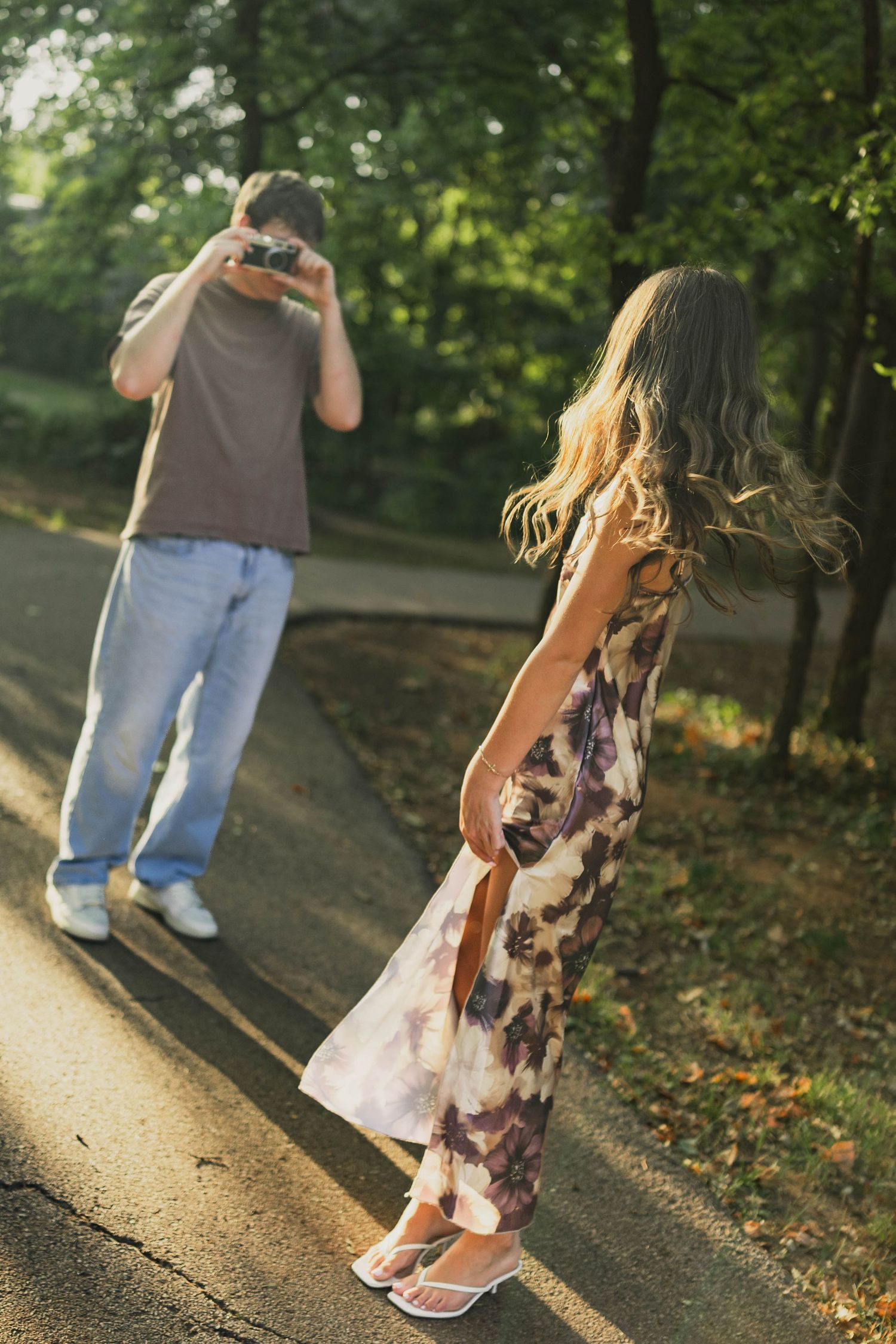 A man photographs a woman in a floral dress on a sunlit park path surrounded by lush green trees.