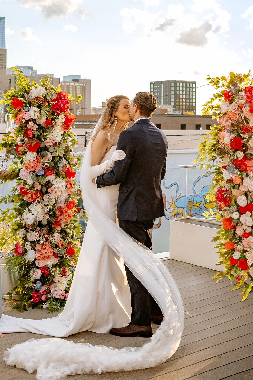 Bride and groom share first kiss on rooftop venue surrounded by vibrant pink and red floral arch with city skyline backdrop.