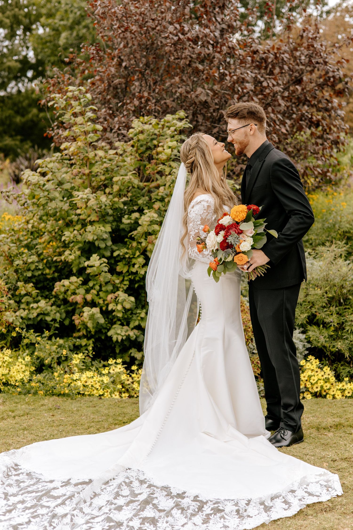 Bride and groom share romantic almost-kiss moment in lush garden, brides lace train spread on grass.