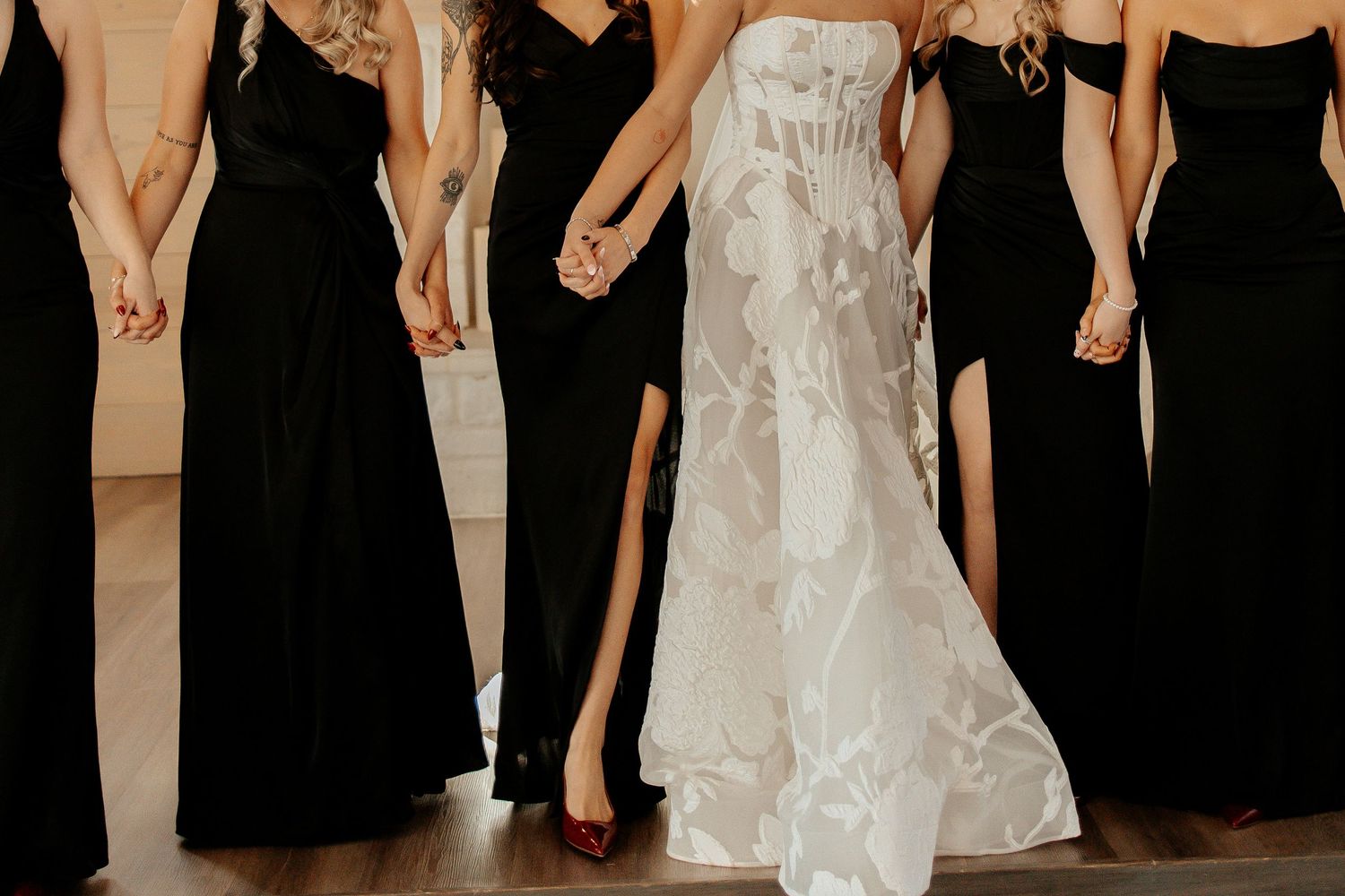 Bride in white floral gown holding hands with bridesmaids in black dresses at wedding ceremony.