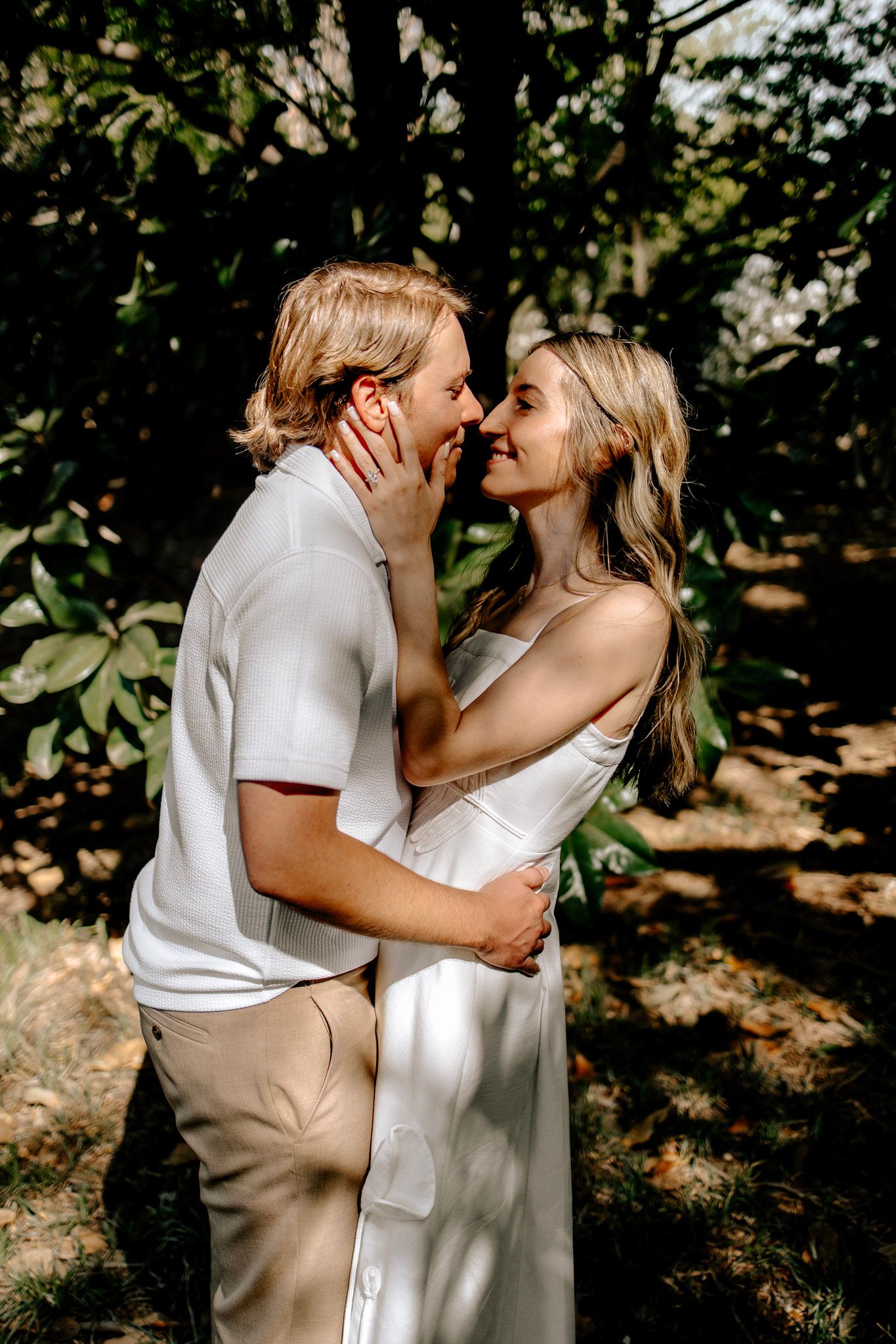 Couple embracing and about to kiss in dappled shade beneath lush green trees, both in light neutral tones.
