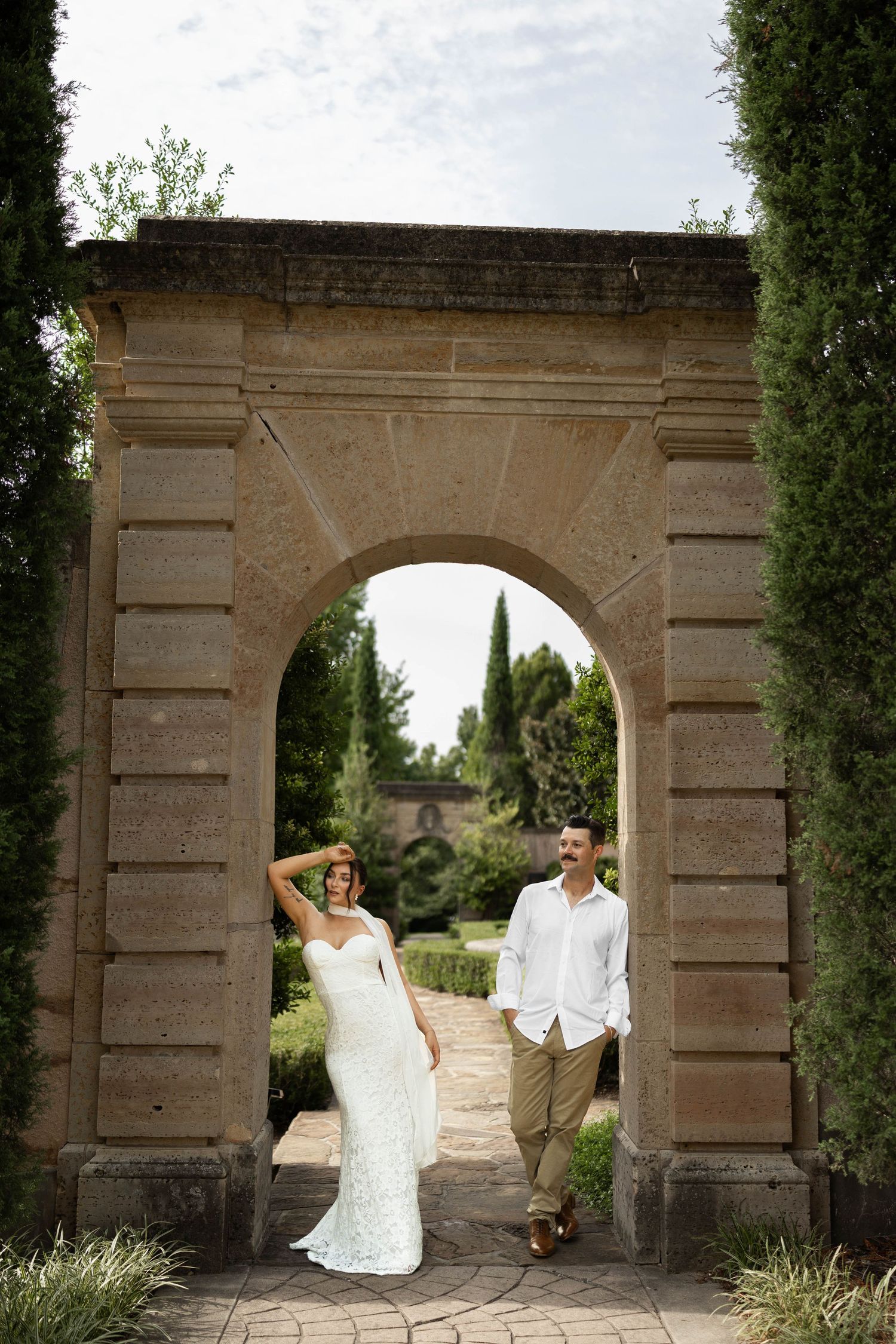 Bride in white gown and groom in white shirt pose under stone arch in lush garden wedding photo.