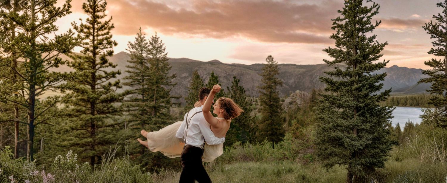 Couple embracing at sunset in a mountain forest, groom lifting bride in wedding attire with pine trees and lake backdrop.
