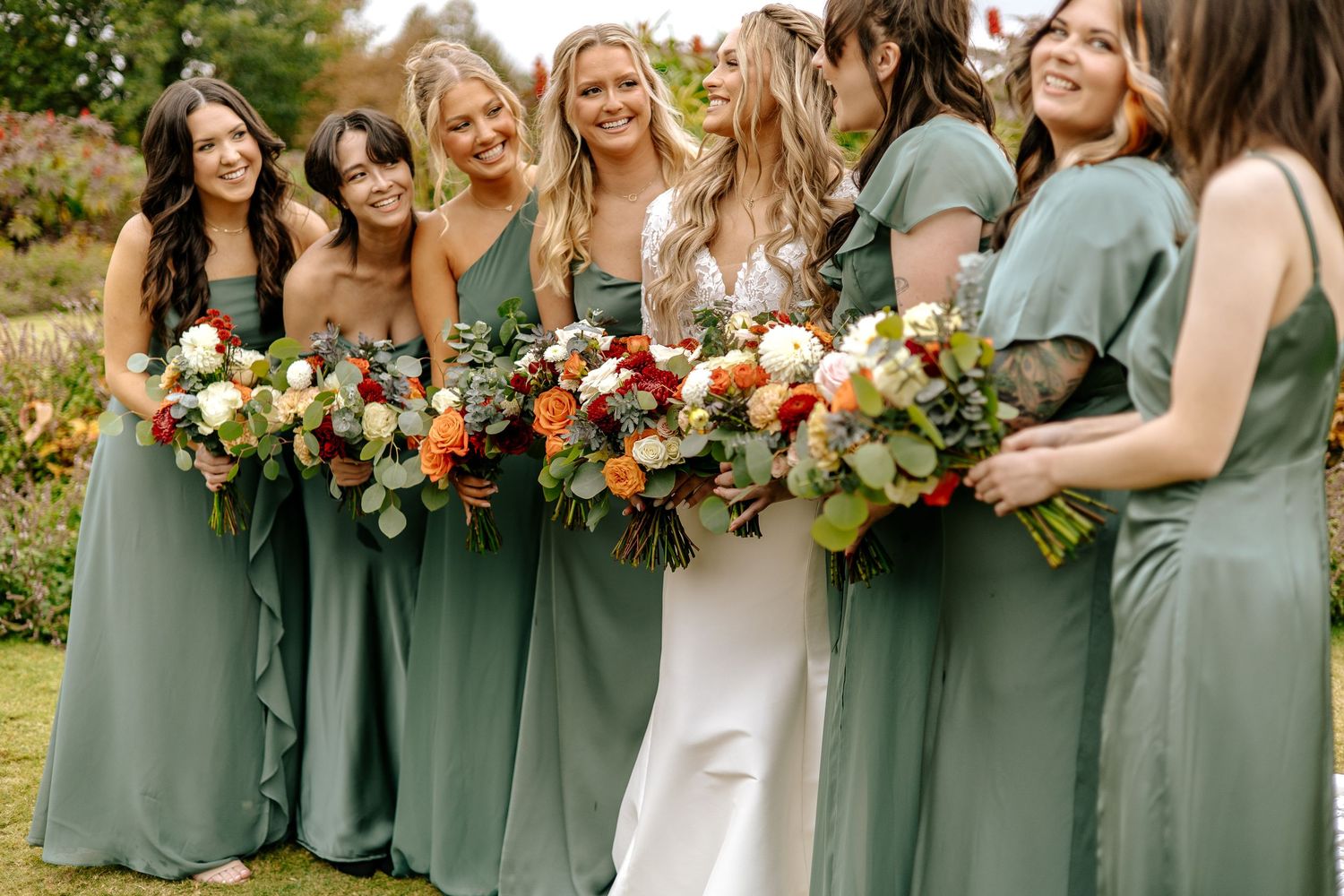 Smiling bride and bridesmaids in sage green dresses laugh together outdoors, holding stunning fall floral bouquets.