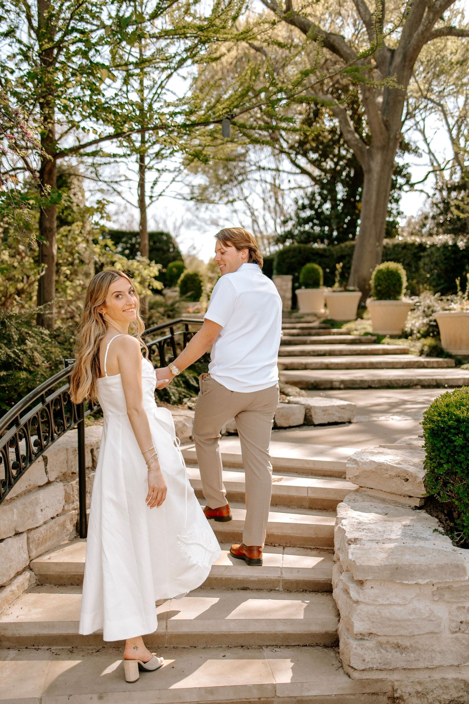 Couple holding hands on stone garden steps, woman in white midi dress glancing back, man in white polo leading the way.