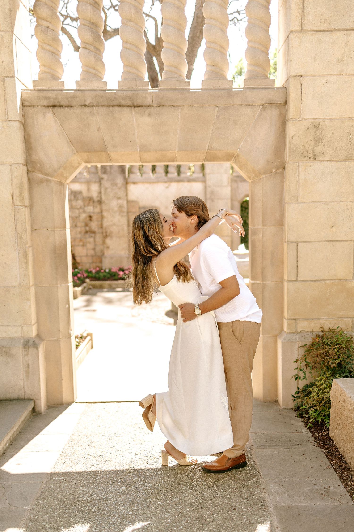 Couple sharing a romantic kiss under a stone archway in a sunlit garden during their engagement session.