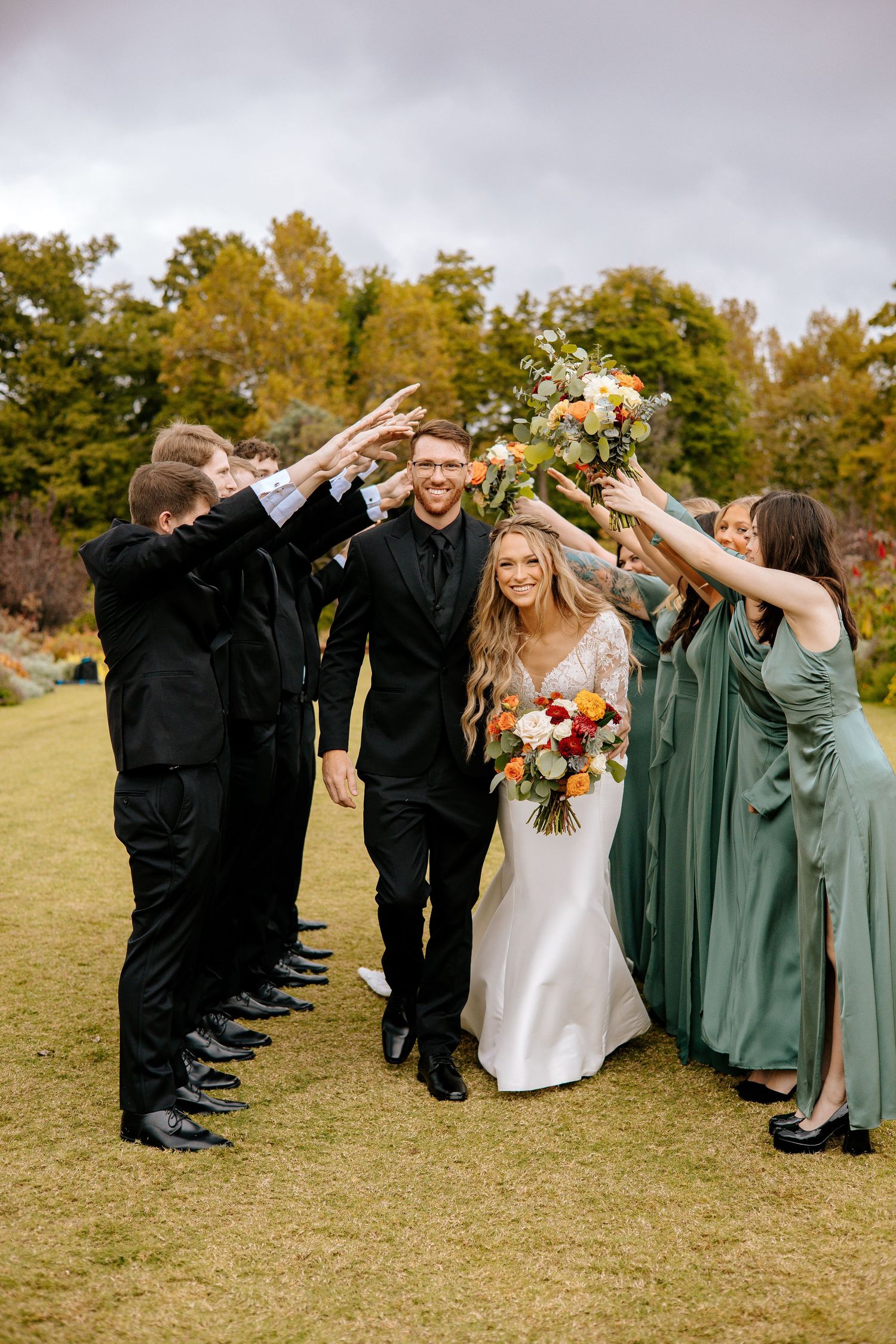 Bride and groom walk through tunnel of raised hands formed by wedding party in black suits and sage green bridesmaid dresses.