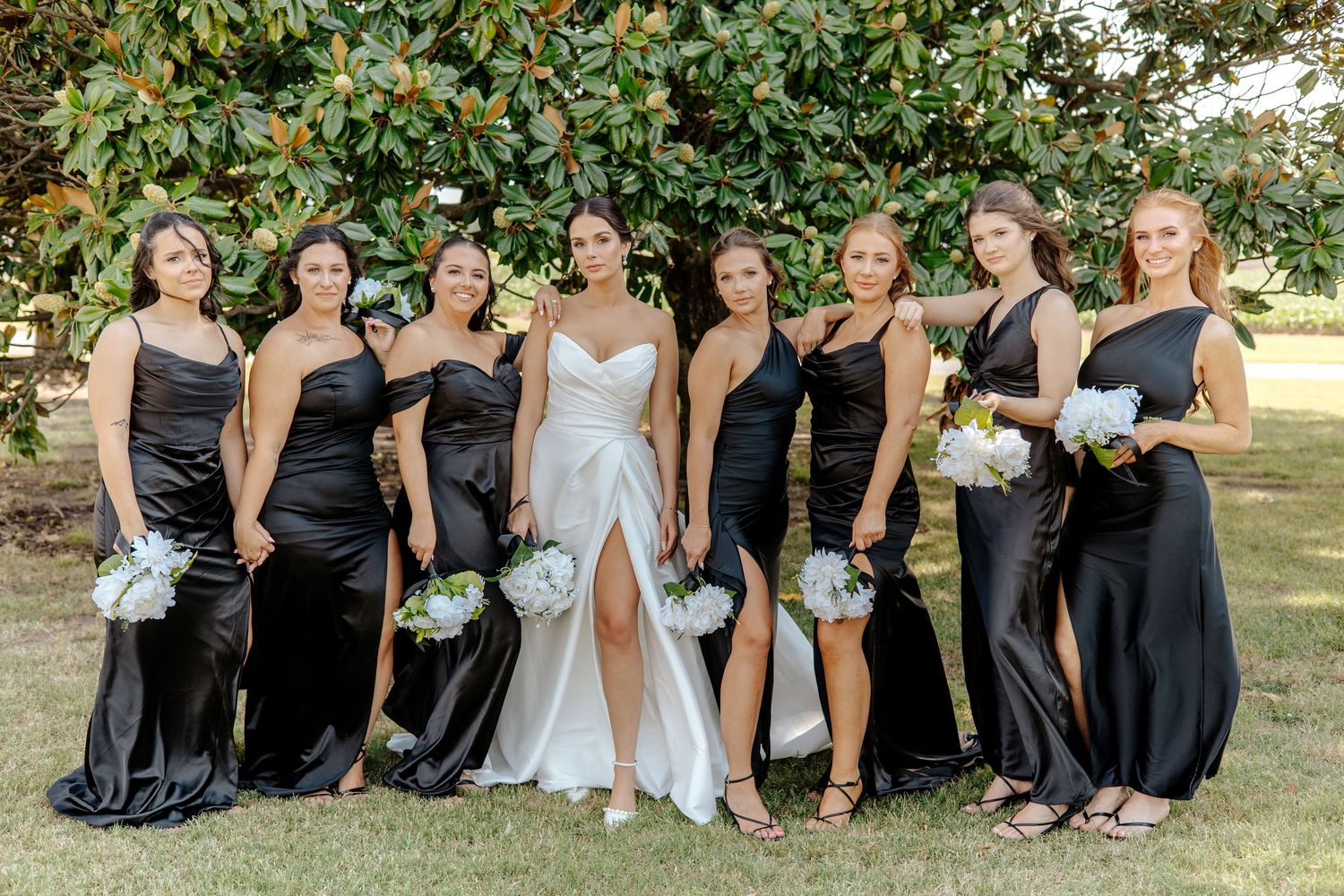 Bride in white gown poses with seven bridesmaids in black satin dresses holding white bouquets.