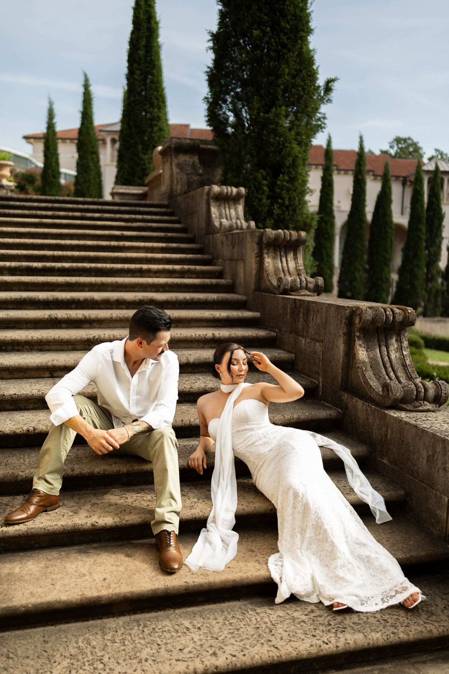 Bride in white gown and groom in white shirt sit on stone steps of elegant garden with cypress trees.