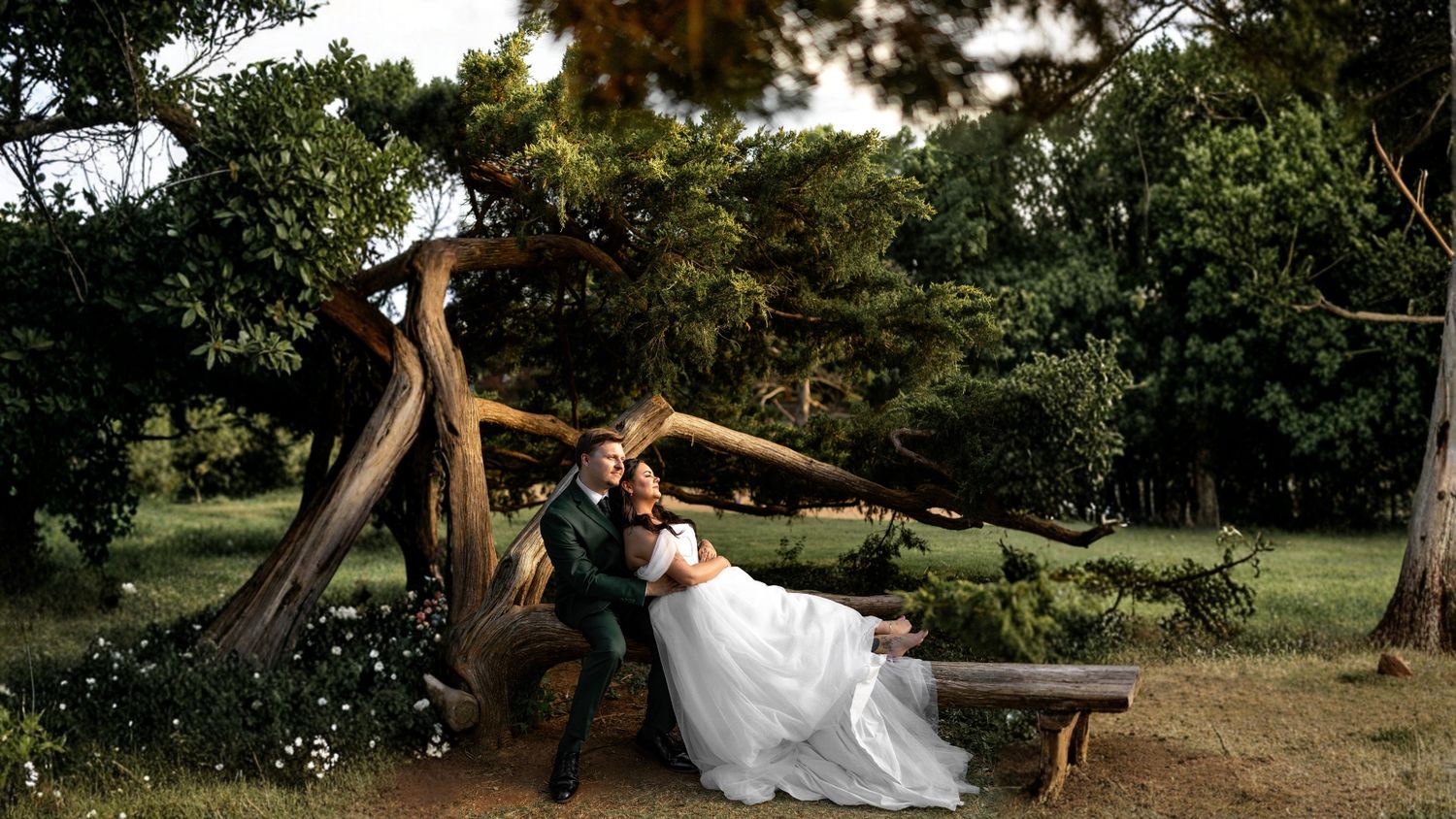 Bride and groom share romantic moment leaning against gnarled tree trunk in lush garden setting during outdoor wedding photo session.