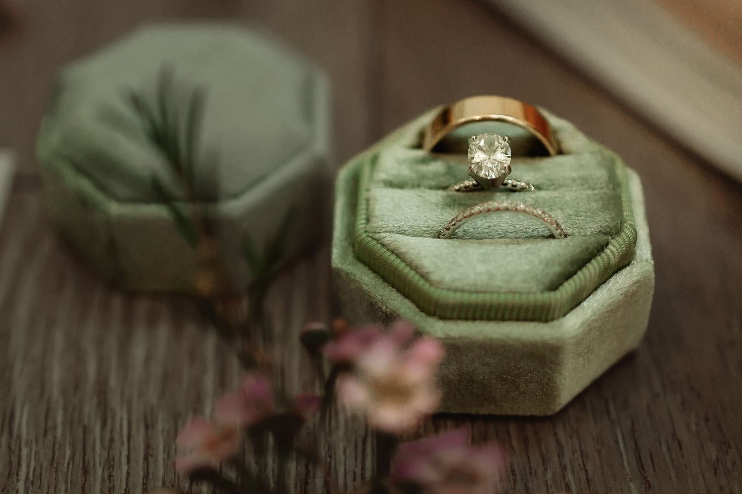 Diamond engagement ring and gold wedding band displayed in a green velvet octagonal ring box on a wooden surface.