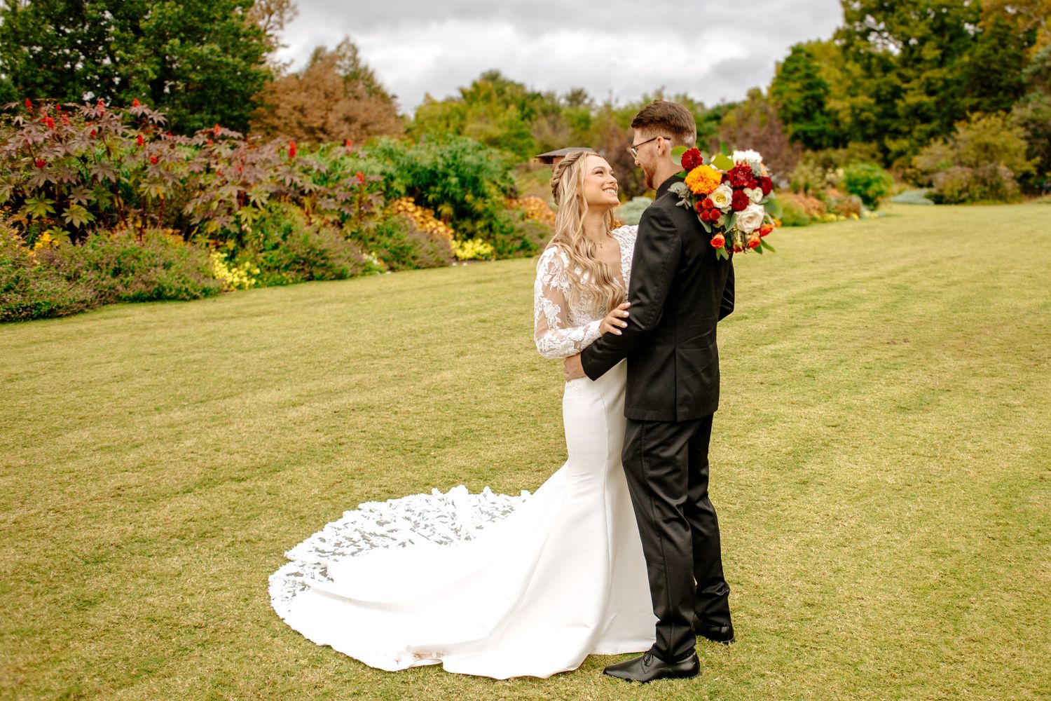 Bride in lace gown holding vibrant bouquet embraces groom in black tuxedo in colorful garden.