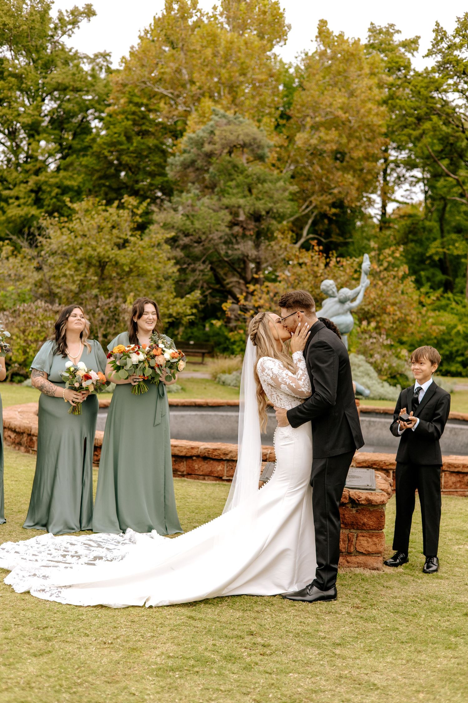 Bride and groom share first kiss at outdoor garden wedding ceremony with bridesmaids in sage green dresses nearby.