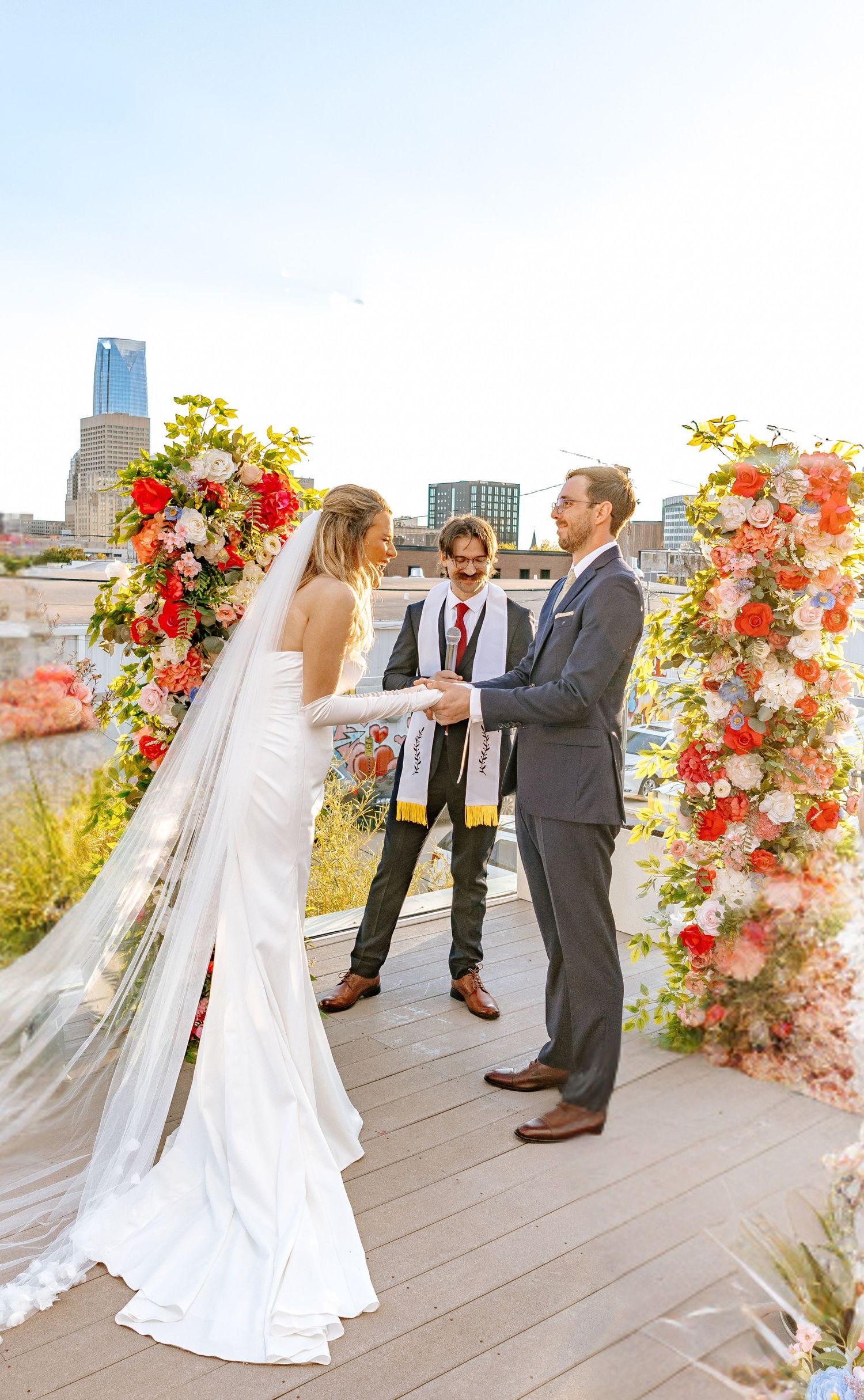 Bride and groom exchange vows at floral arch on rooftop venue with city skyline backdrop on sunny wedding day.