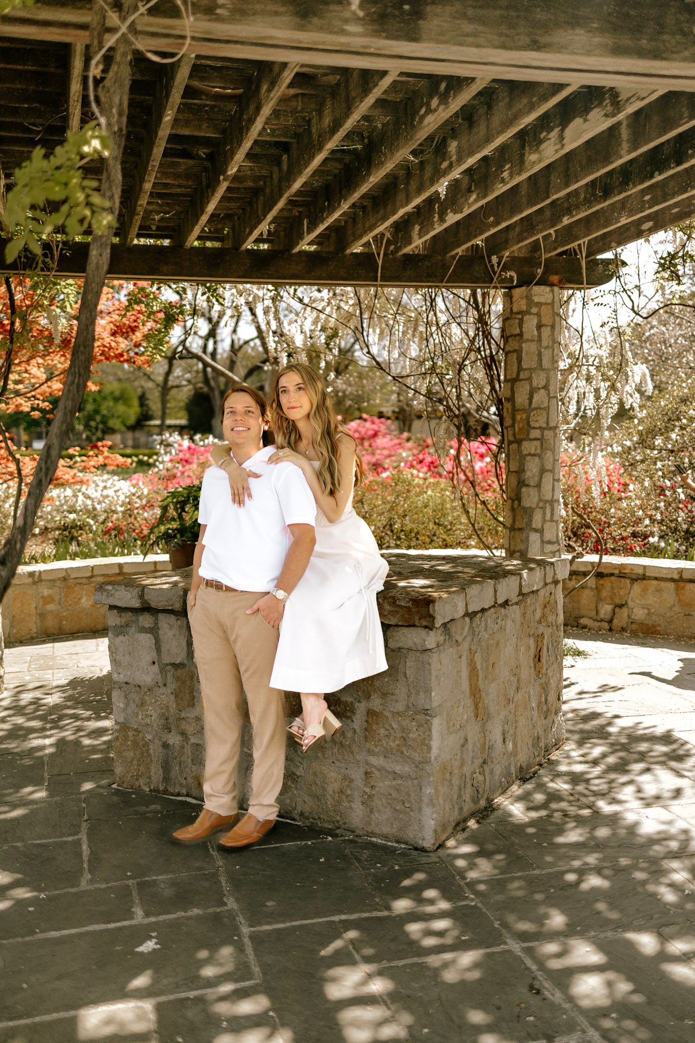 Couple standing under a wooden pergola surrounded by vibrant fall foliage in a garden setting.