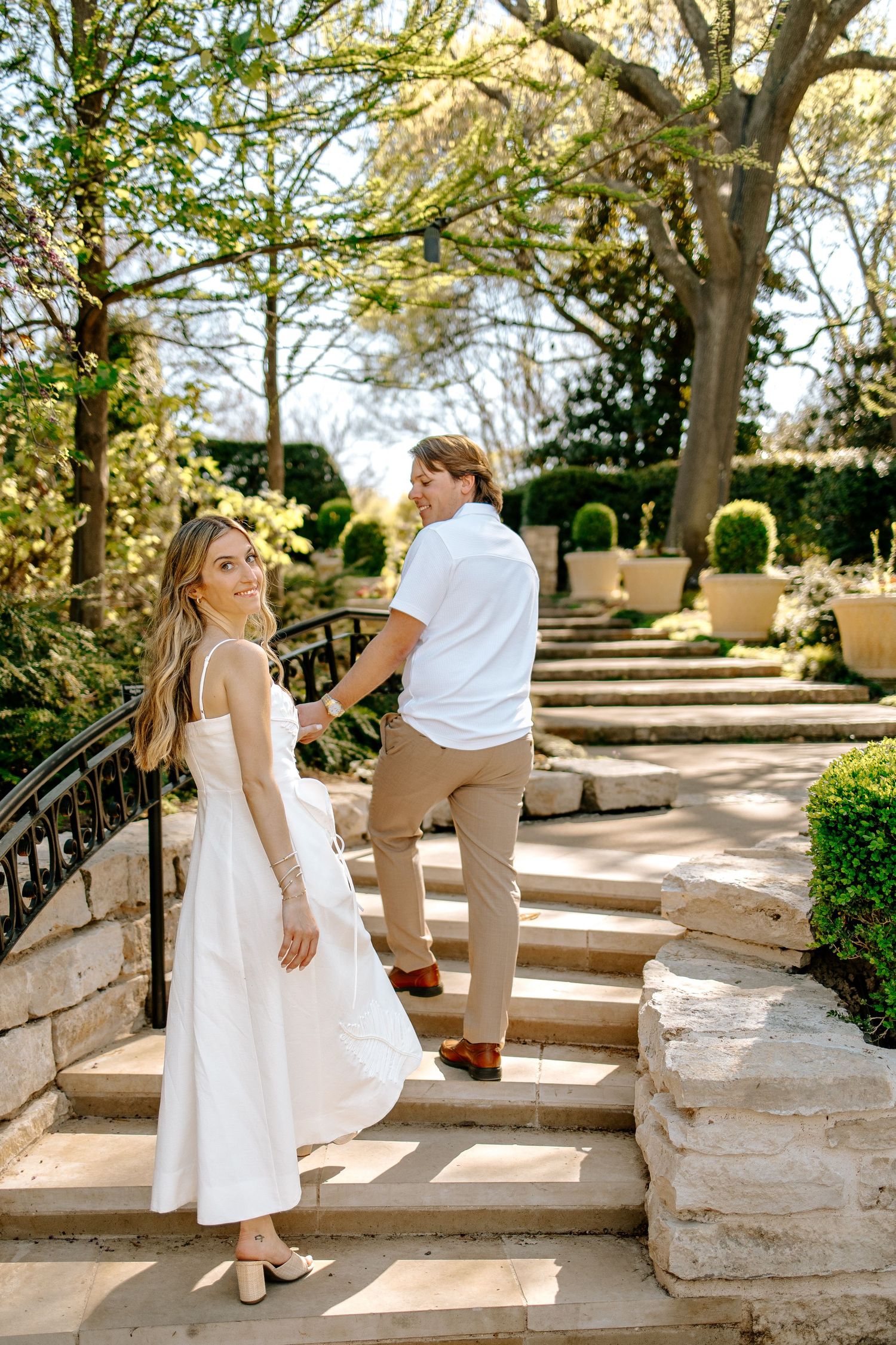 Bride in white dress and groom in white shirt hold hands ascending stone garden steps.