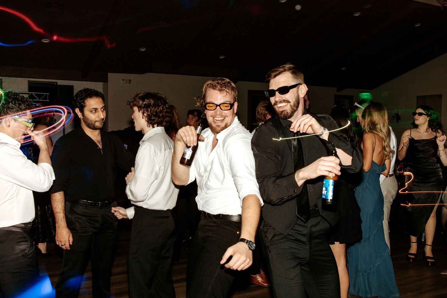 People dancing and posing at a party with neon glow sticks, wearing formal attire and sunglasses on a dance floor.