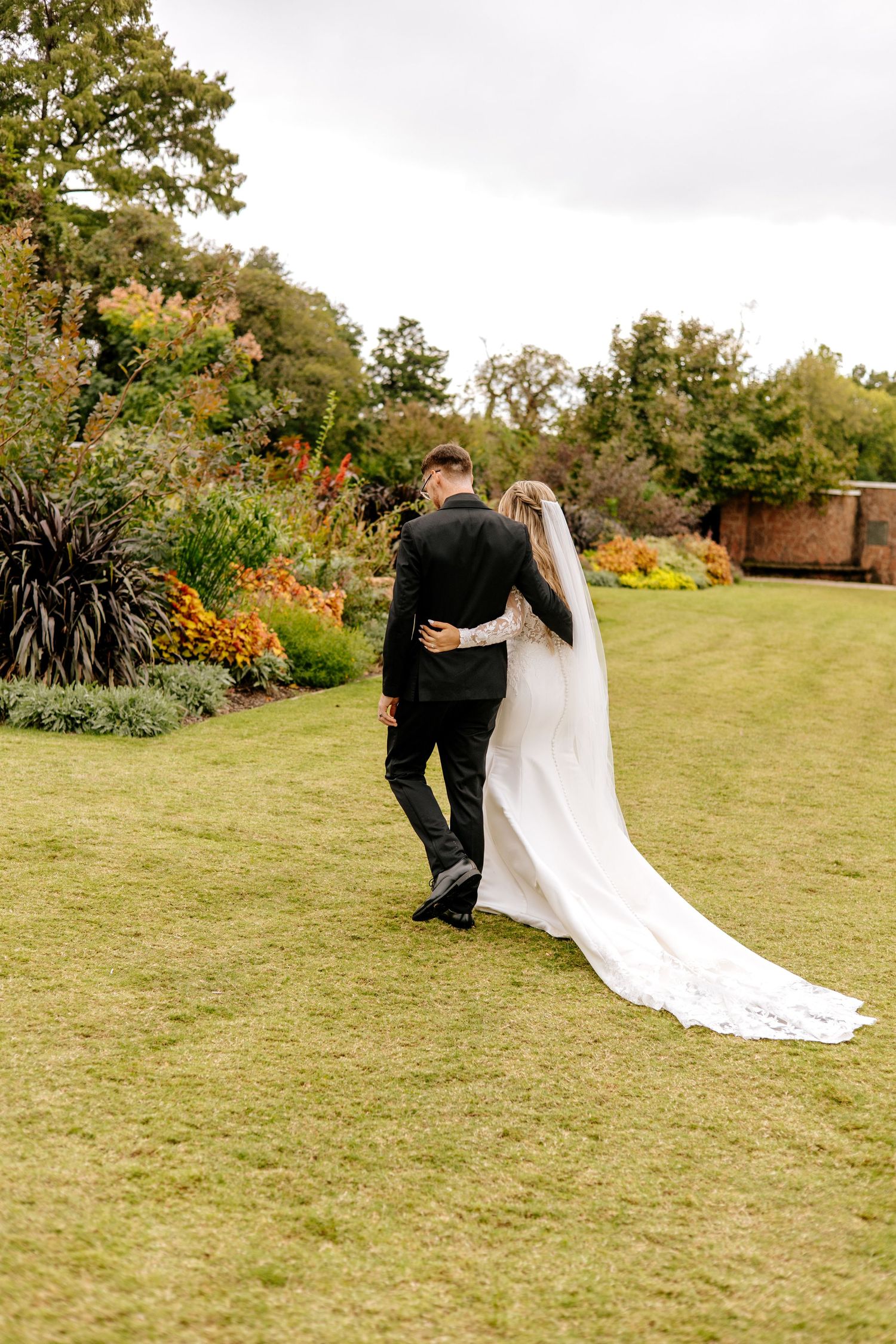 Newlyweds walk arm in arm through colorful autumn garden, bride in fitted white gown with long cathedral veil trailing.