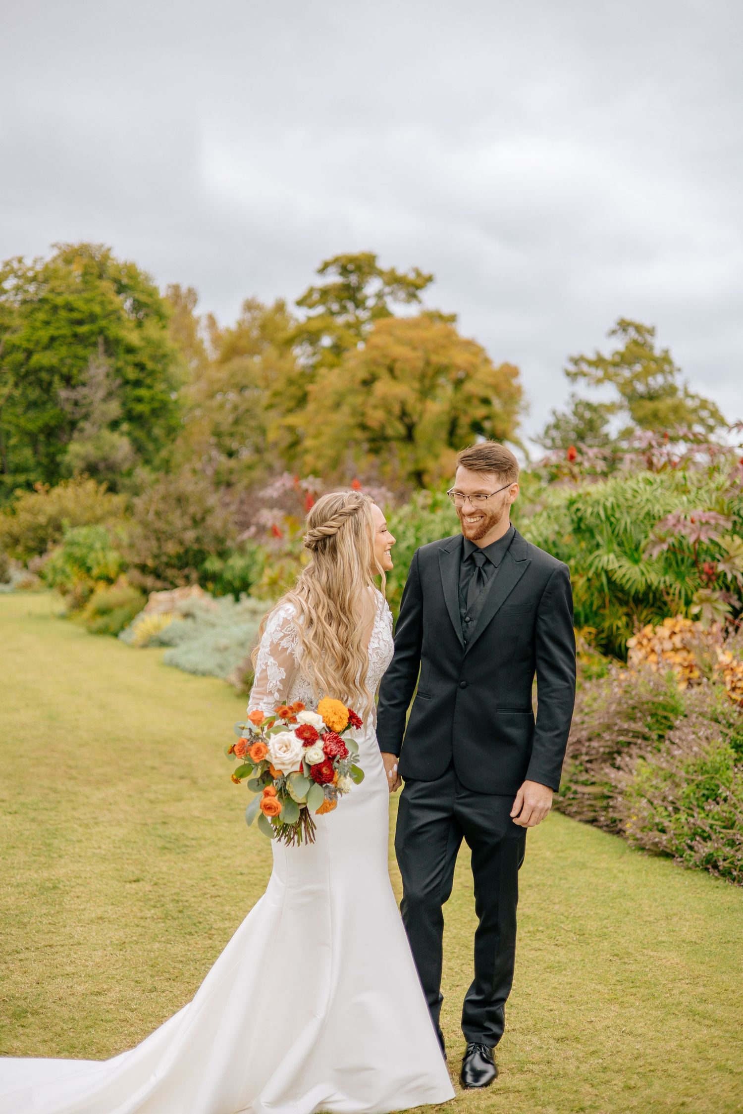 Bride and groom walk hand-in-hand through a lush garden, bride holding a vibrant autumn bouquet in a lace wedding gown.