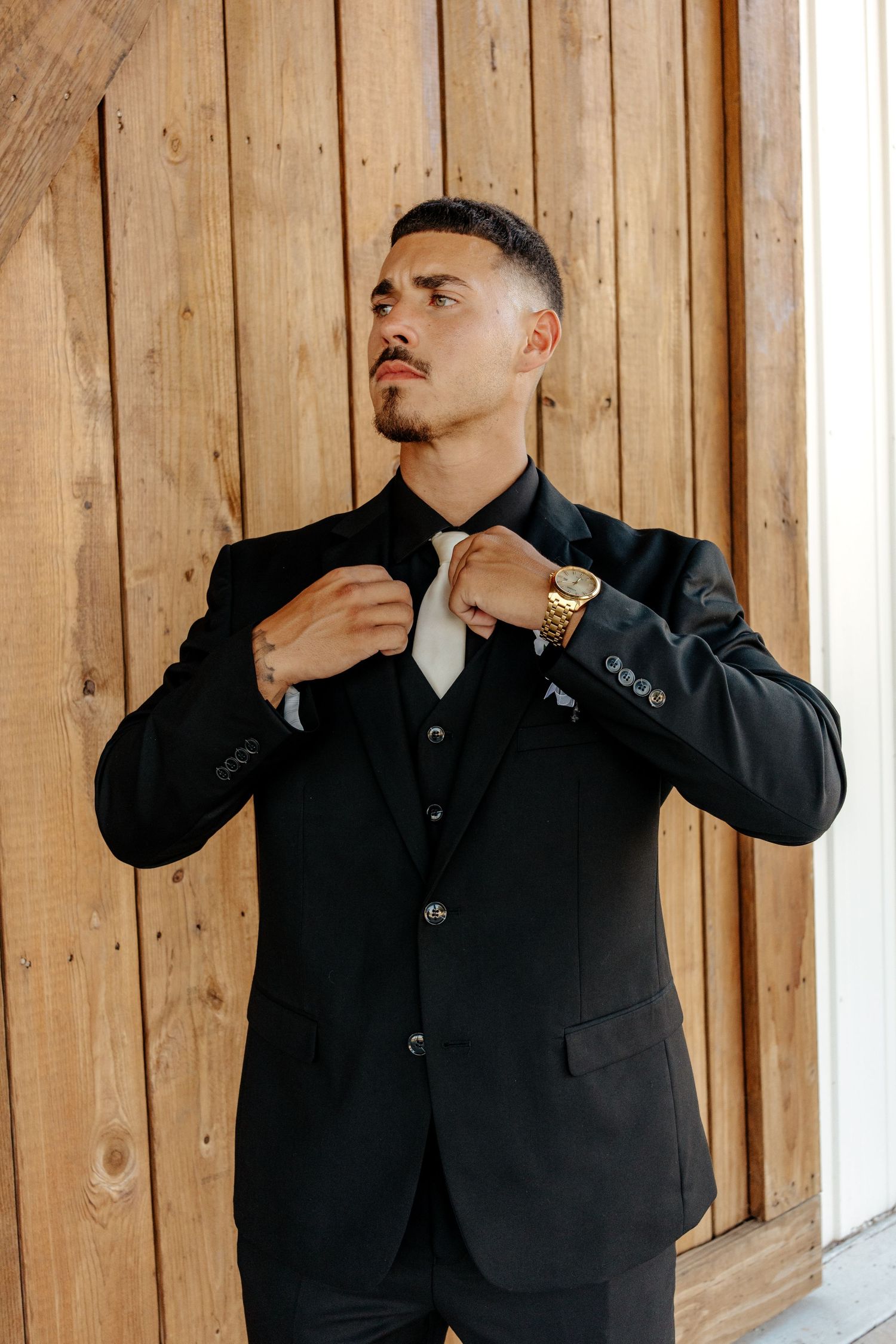 Handsome groom straightening white tie in black suit with gold watch at barn venue.