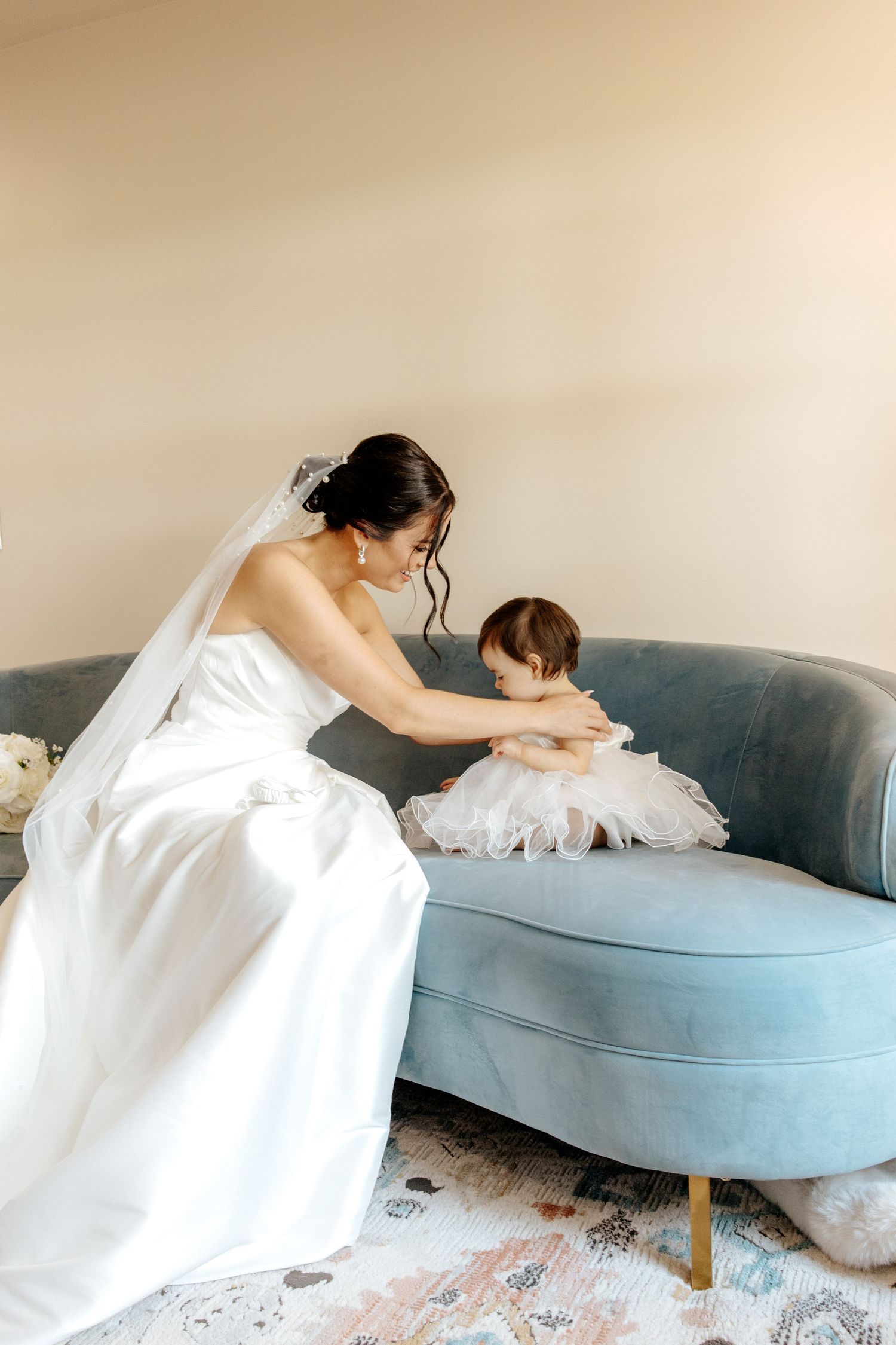 Bride in white wedding gown and veil interacts with baby flower girl on blue velvet sofa.