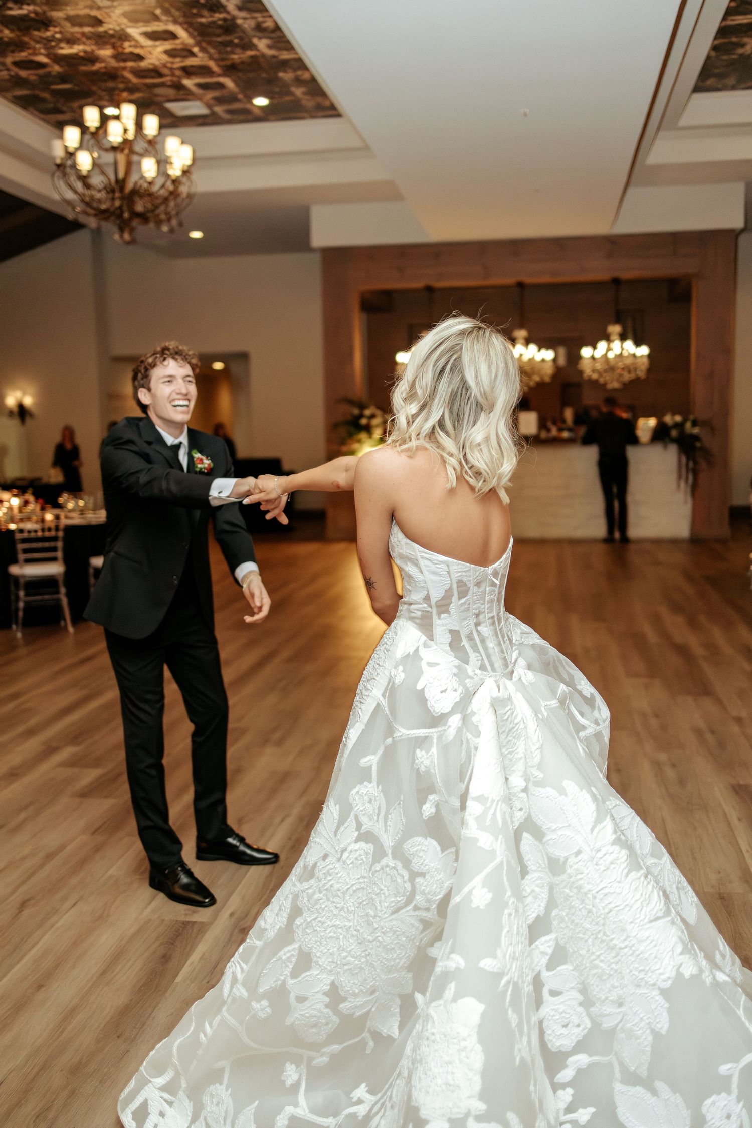 Smiling groom spins bride in elaborate floral ballgown during first dance at wedding reception.