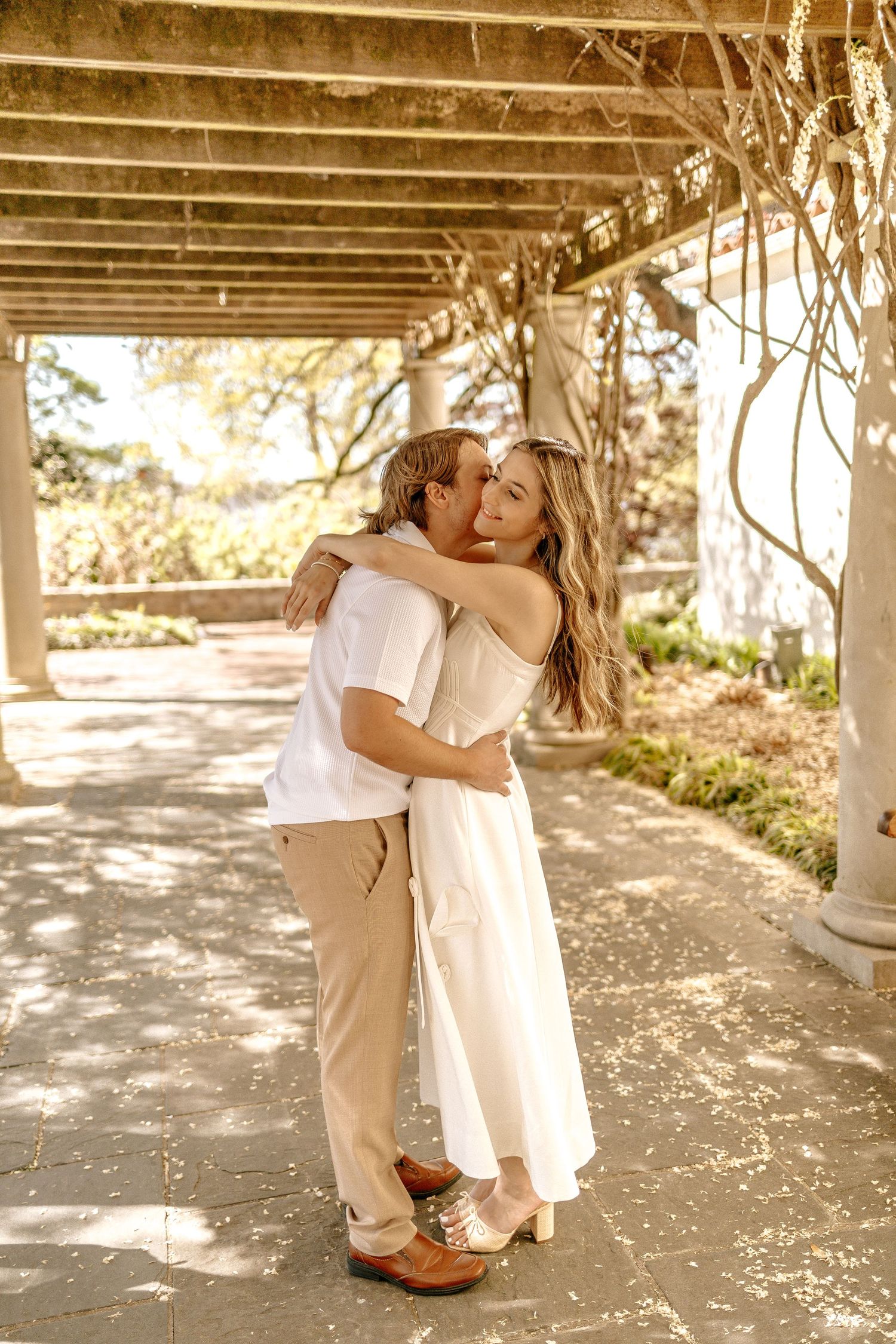 Couple embracing and kissing under a vine-covered pergola in a sun-dappled garden pathway.