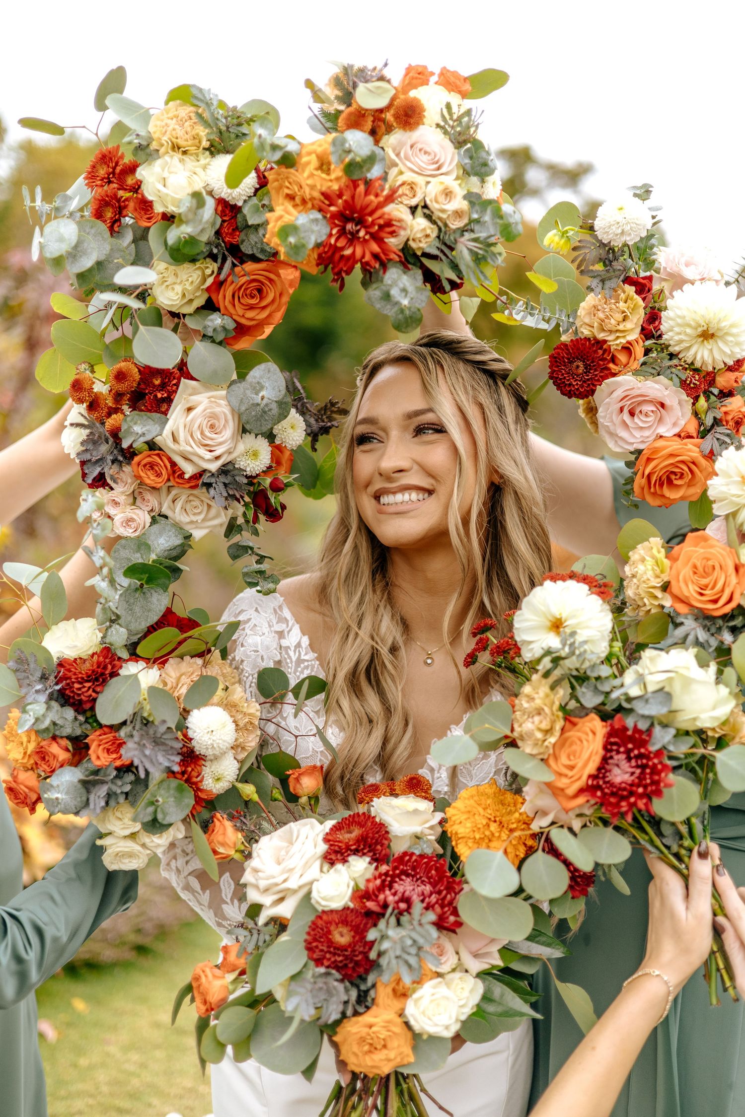 Smiling bride framed by a stunning circular floral arrangement of orange, red, and white autumn flowers and eucalyptus.