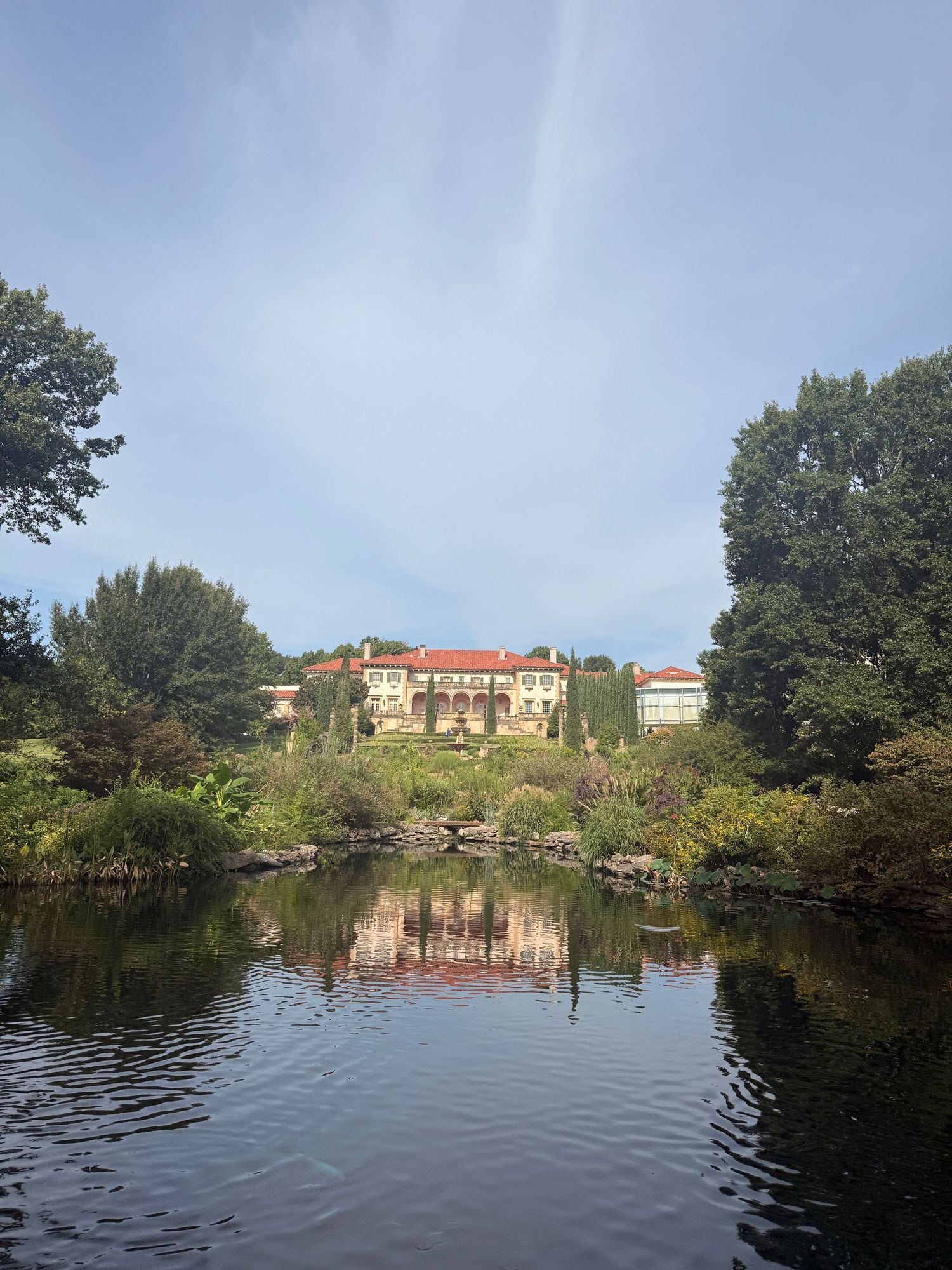 A grand Italian-style villa reflects in a calm pond surrounded by lush green trees under a blue sky.