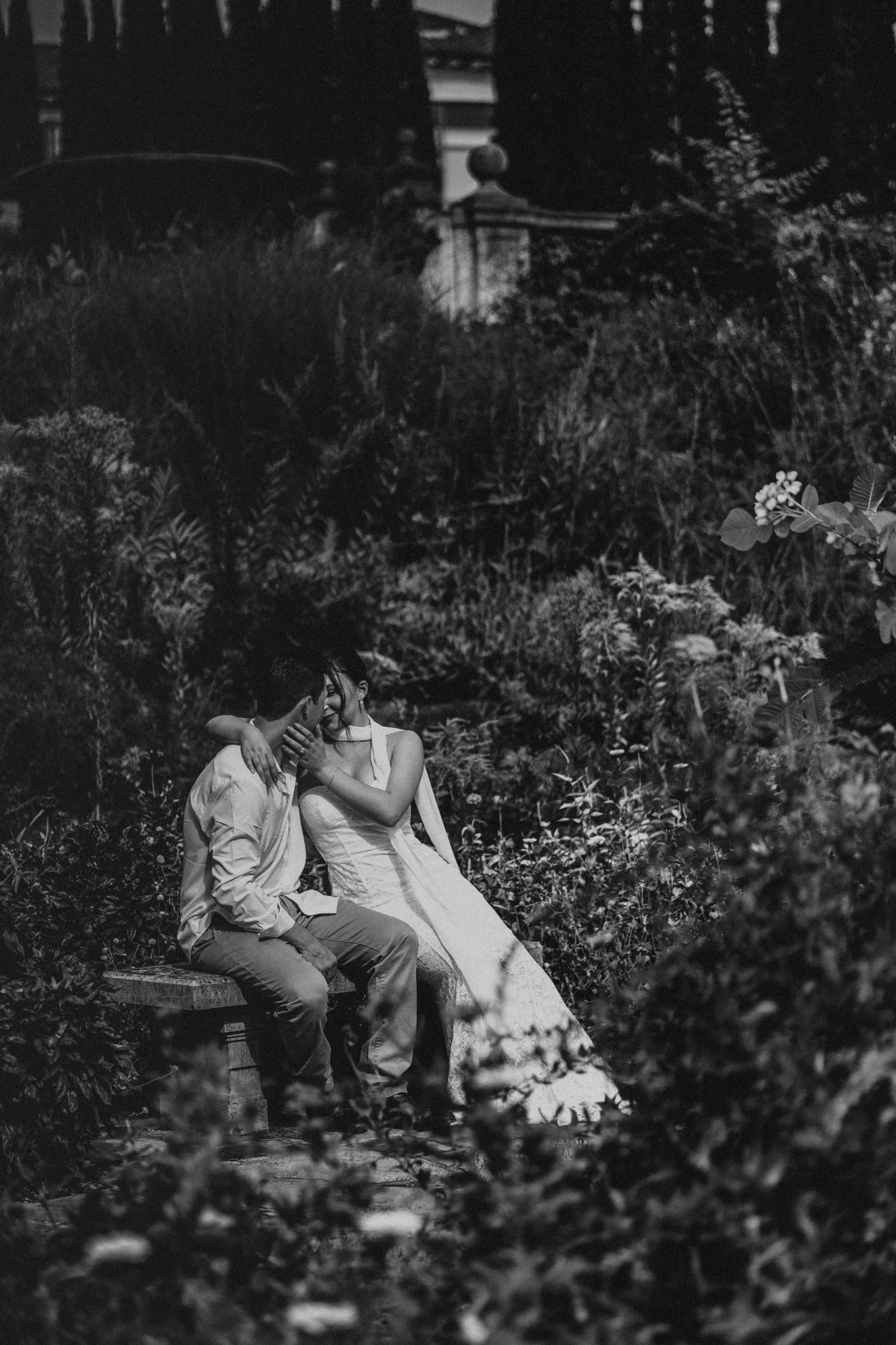 Black and white photo of a bride and groom sitting on a garden bench surrounded by lush flowers and greenery.