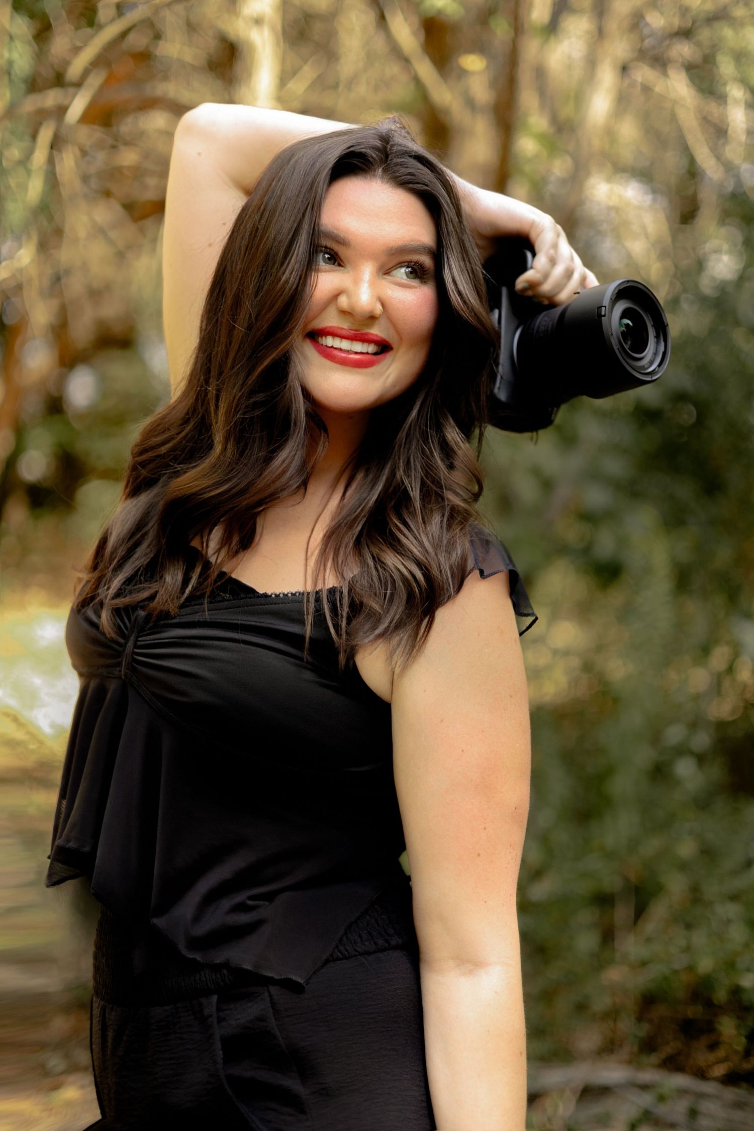 Smiling brunette woman in black outfit holding a camera outdoors in a lush green forest setting.