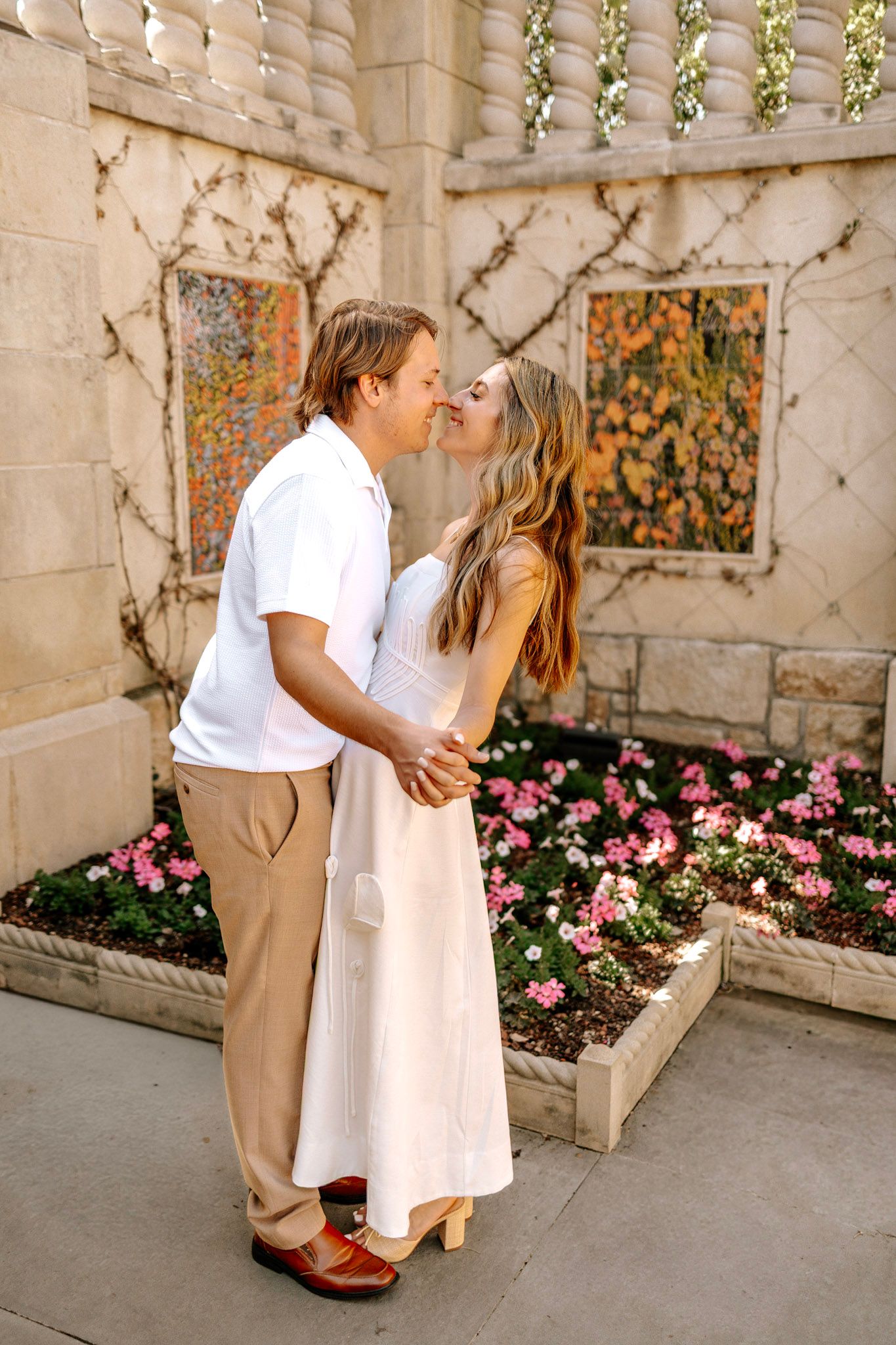 Couple kissing in front of a stone wall with pink flowers, woman in white dress holding hands with man.