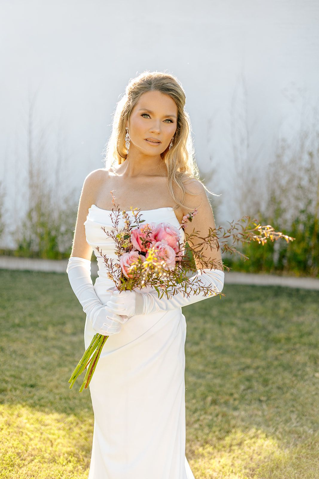 Elegant blonde bride in strapless white gown and gloves holding a pink and yellow bridal bouquet in sunlit outdoor setting.